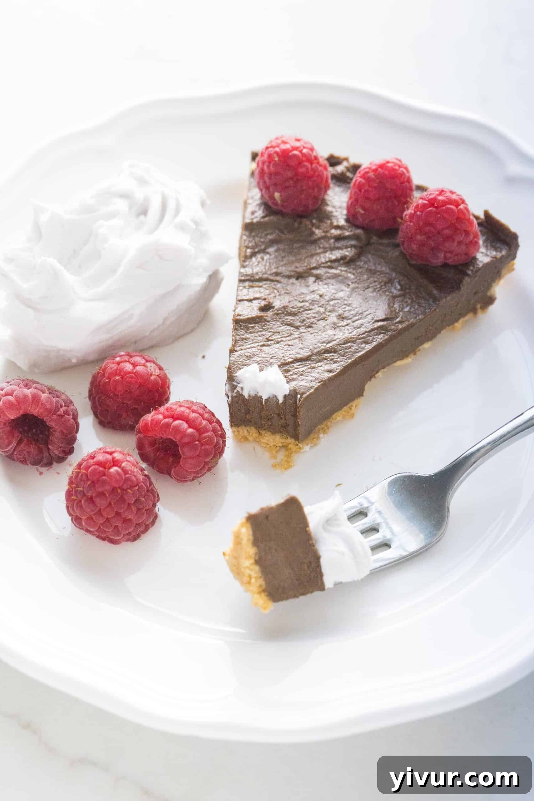 A fork taking a delicious bite out of a healthy chocolate tart, garnished with vibrant raspberries and fluffy coconut whipped cream.