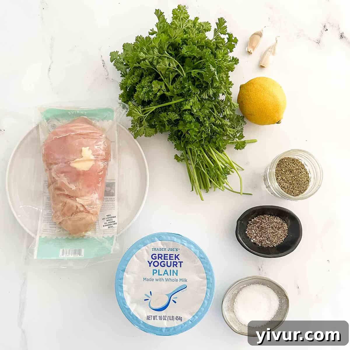 Overhead view of recipe ingredients for Greek yogurt chicken arranged on a white surface, including Greek yogurt, fresh garlic cloves, a whole lemon, dried herbs, and raw chicken pieces.