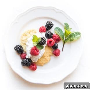 A keto berry shortcake biscuit topped with fluffy whipped cream, fresh raspberries, blackberries, and a sprig of mint on a white plate.