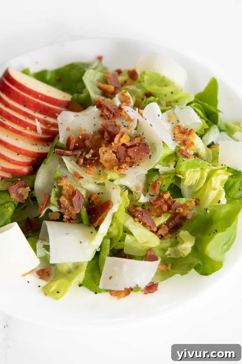 A fresh Butter Leaf Lettuce Salad topped with crisp apple slices, crunchy bacon bits, and delicate shaved parmesan, served on a pristine white plate.