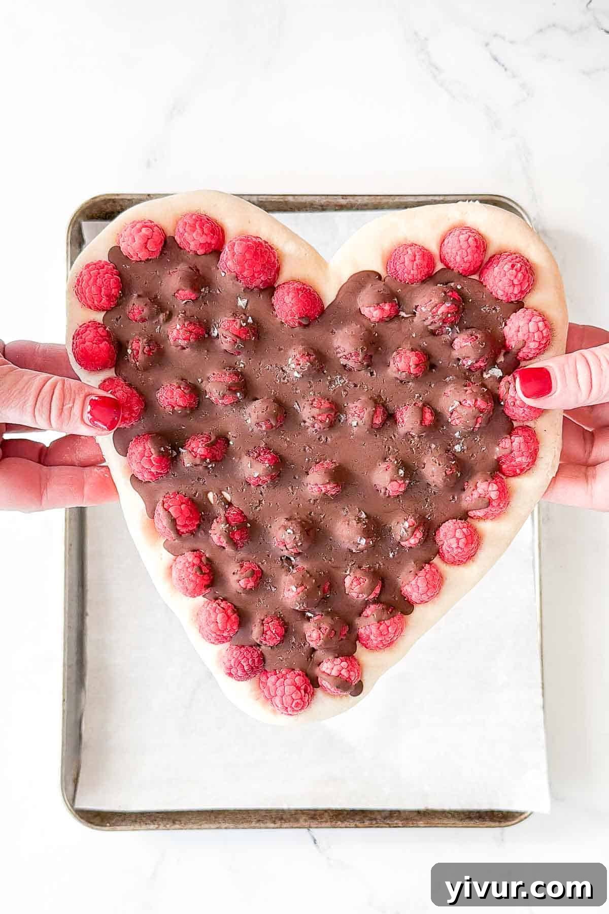 A top-down view of vibrant pink yogurt bark shaped like a heart, studded with fresh red raspberries and drizzled with melted dark chocolate, presented on a white plate.