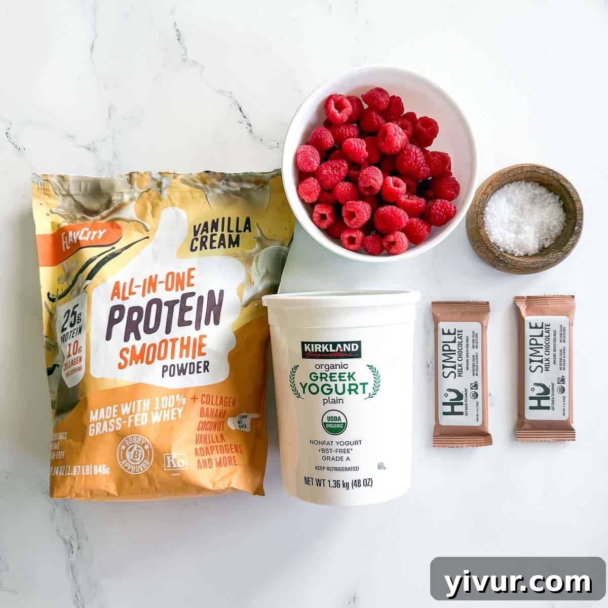Close-up of fresh ingredients laid out: a bowl of Greek yogurt, a scoop of vanilla protein powder, a container of fresh raspberries, a bar of coconut sugar sweetened chocolate, a small spoon of coconut oil, and a pinch of flaky sea salt.