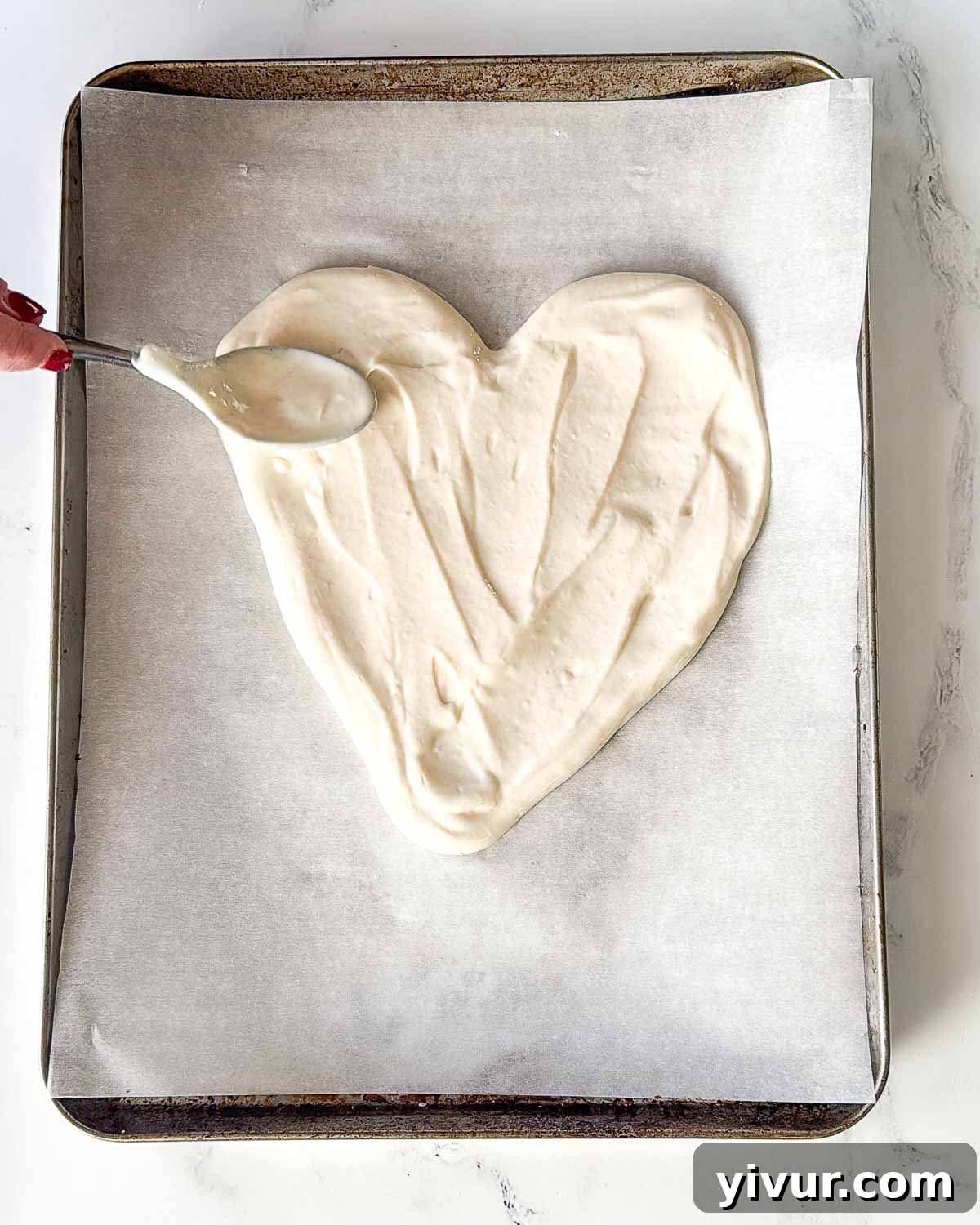 A close-up image showing the creamy Greek yogurt and protein powder mixture being spread evenly onto a parchment-lined baking sheet, carefully shaped into a perfect heart with a spatula.