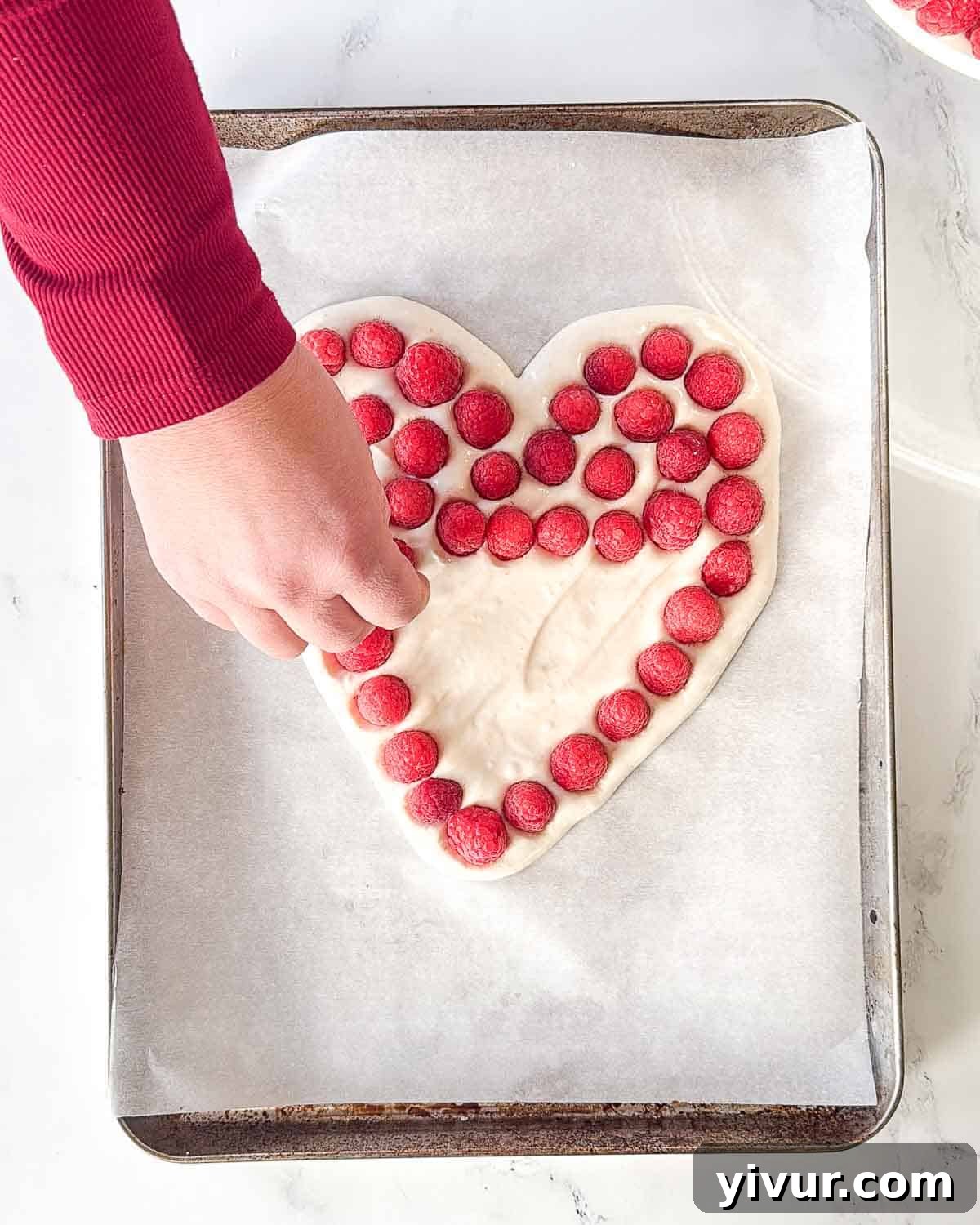 Fresh red raspberries are gently pressed into the surface of the heart-shaped Greek yogurt mixture on a baking sheet, creating a beautiful pattern.