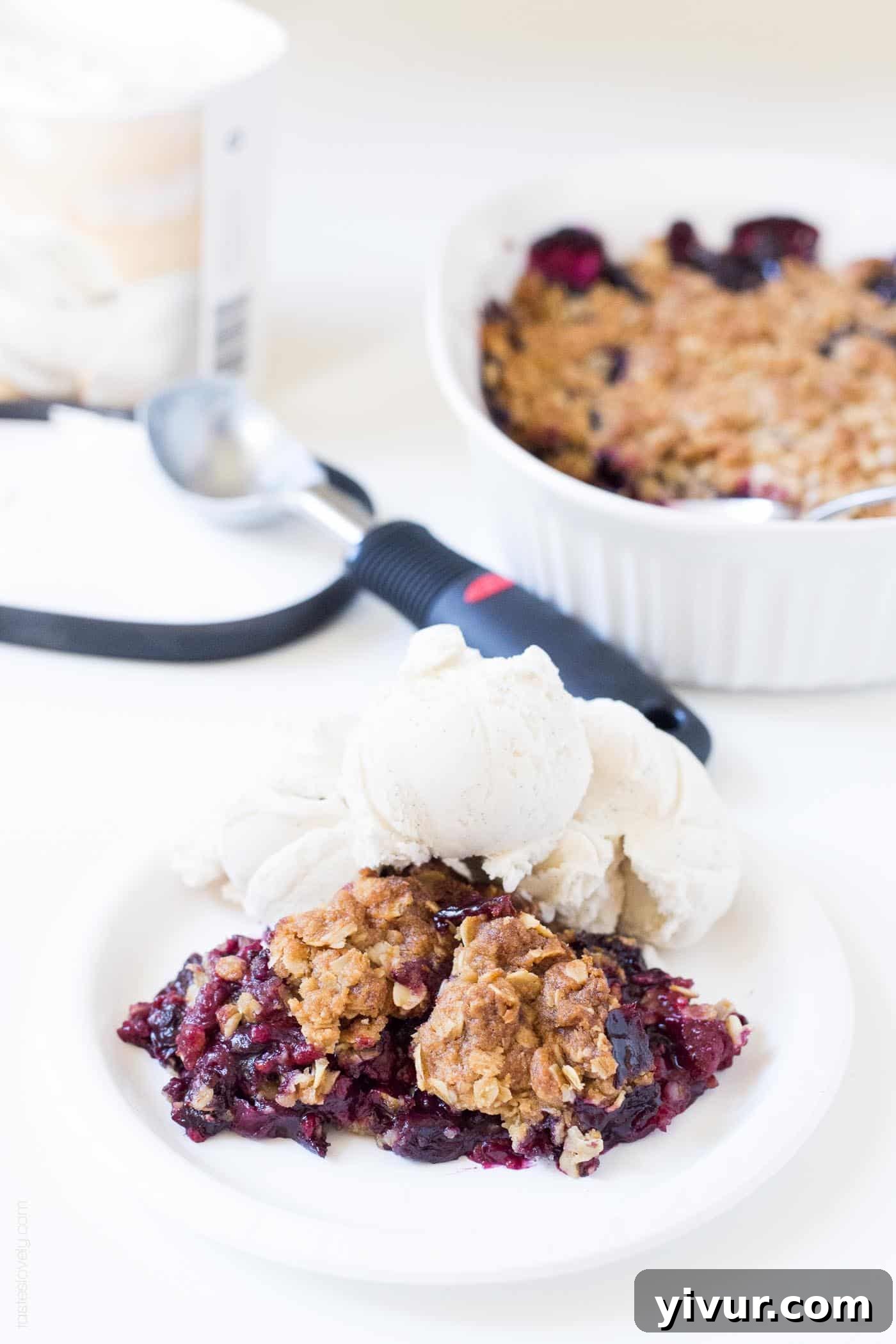 Quick Triple Berry Crumble from Frozen 7 A white plate featuring a slice of triple berry crumble and a scoop of vanilla ice cream, with an ice cream scooper in the background.