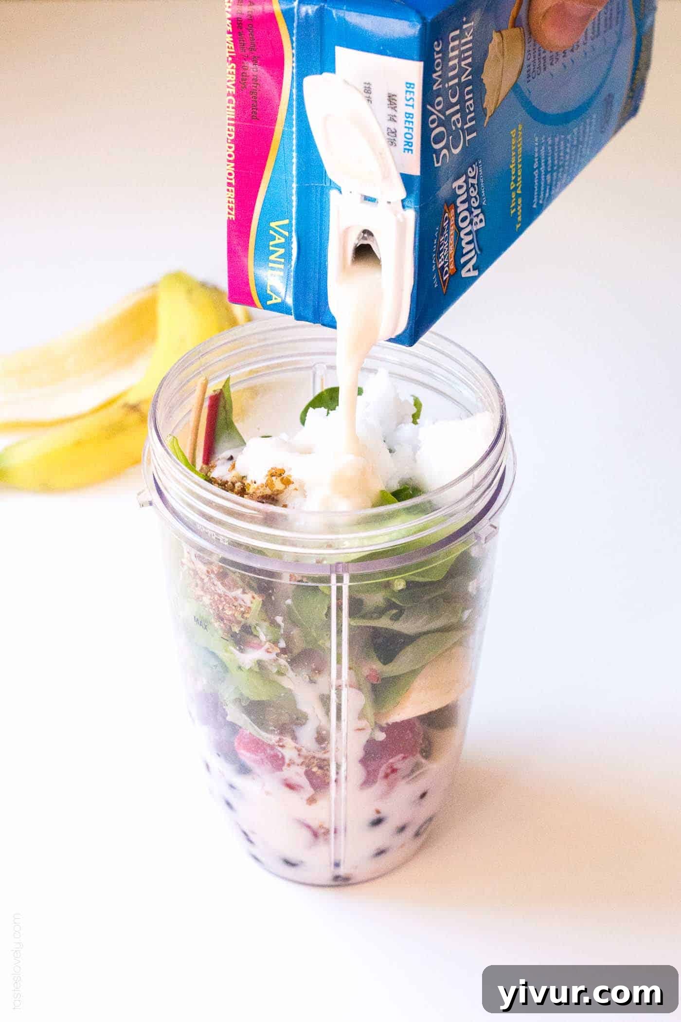 Hand pouring unsweetened almond milk into a blender cup filled with organic fruits and greens, ready to be blended into a healthy pregnancy smoothie.