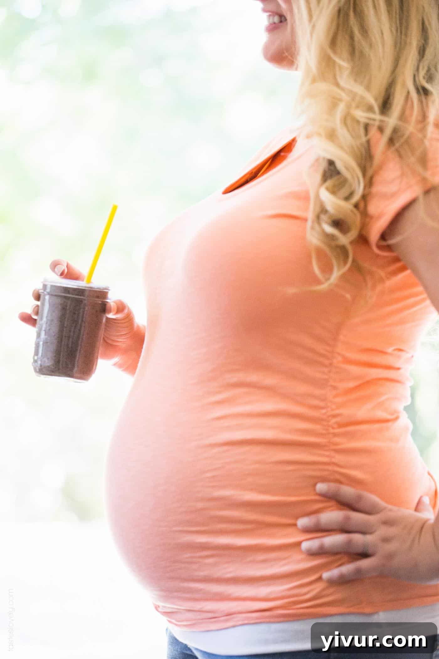 A smiling pregnant woman holding a glass filled with a healthy pregnancy smoothie and a yellow straw, looking refreshed.