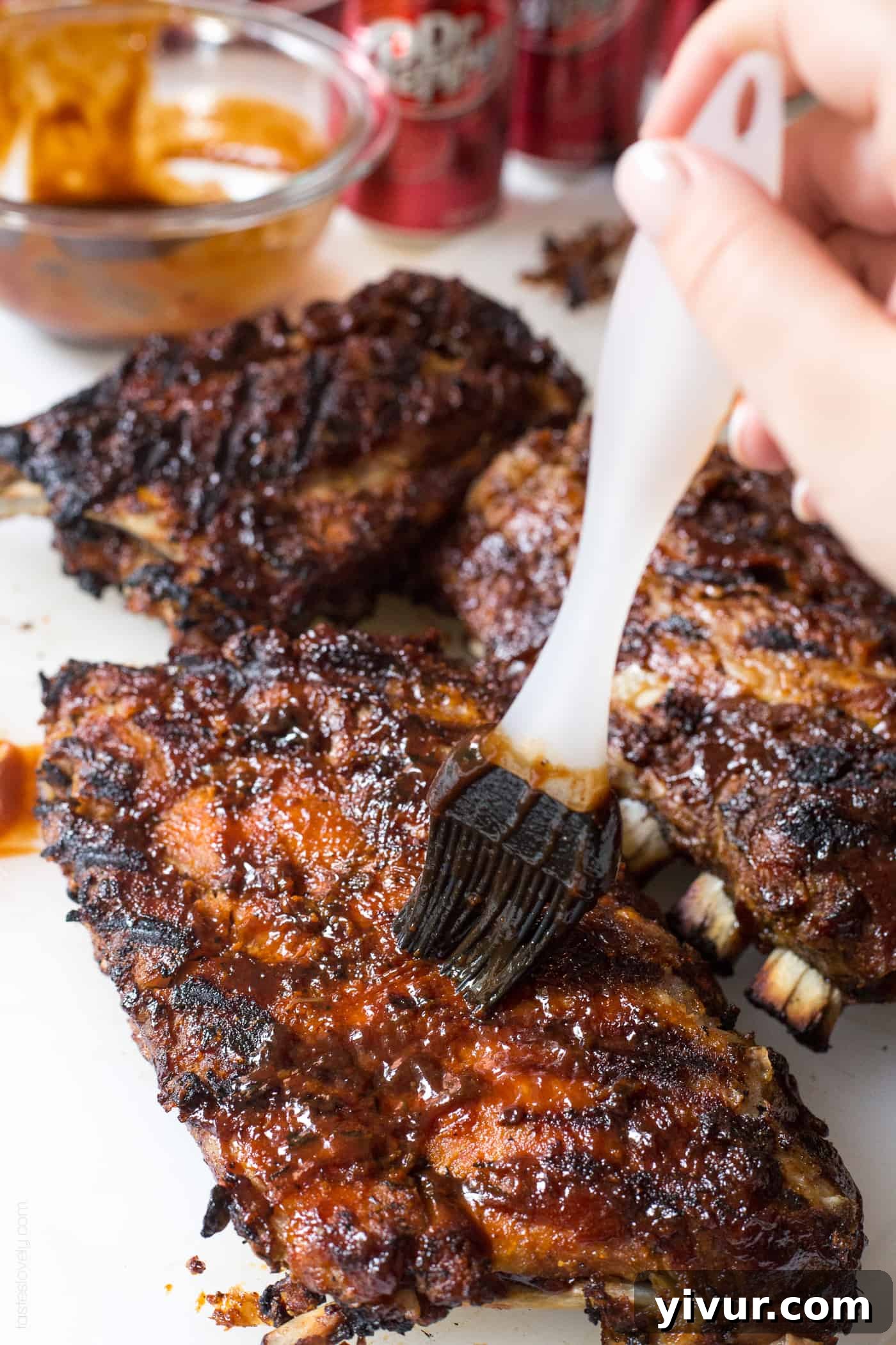 A hand brushing rich BBQ sauce onto a rack of perfectly cooked Dr Pepper ribs on the grill, creating a beautiful glossy finish.
