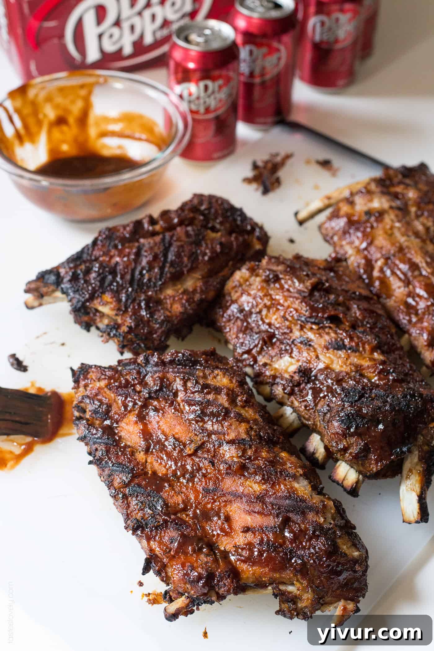 Multiple cans of Dr Pepper arranged on a white surface next to a rack of seasoned Dr Pepper ribs, ready for cooking.