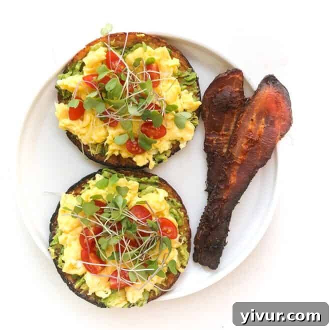 Keto avocado toast topped with a fried egg, ripe tomatoes, microgreens, and crispy bacon on a white plate.