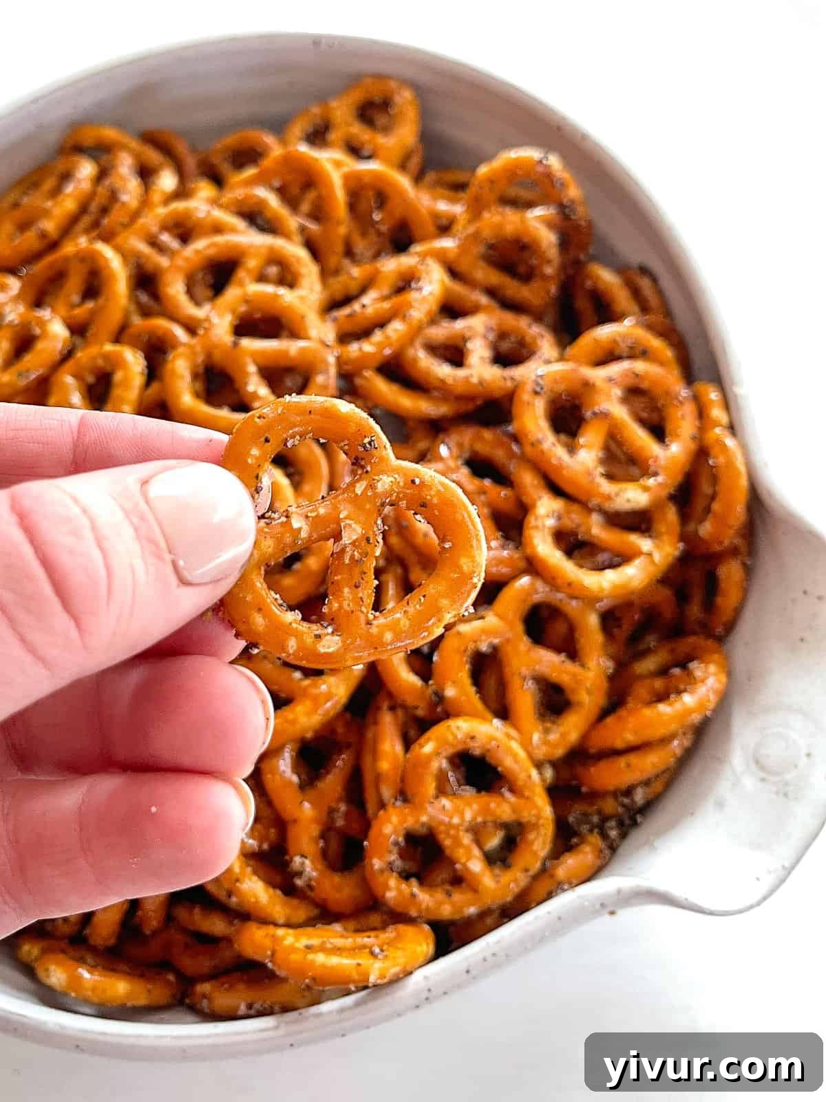 Effortless Fiery Pretzel Crunch 5 A close-up photo of a white bowl filled with pretzel twists, with a hand holding one pretzel close to the camera, emphasizing its seasoning.