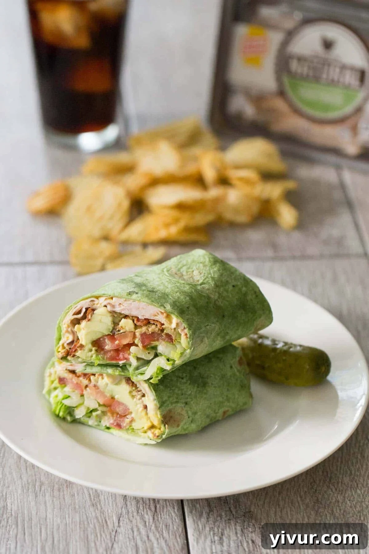 A spinach wrap filled with turkey, lettuce, tomatoes, and cheese is cut in half and placed on a white plate with a pickle. In the background are potato chips and a glass of dark soda.