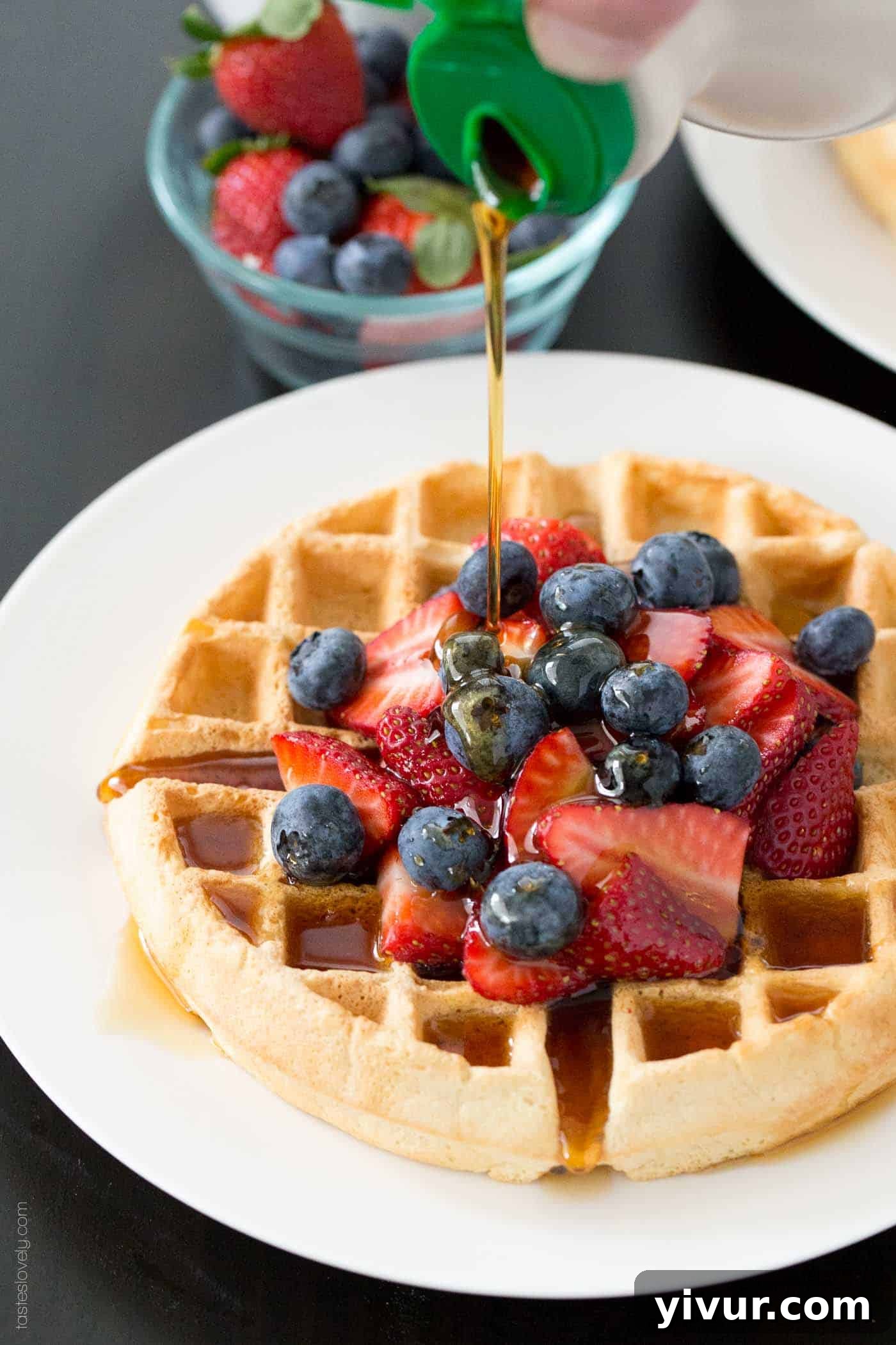 Pure maple syrup being generously poured over a stack of golden-brown dairy-free waffles, adorned with a medley of fresh, colorful berries.
