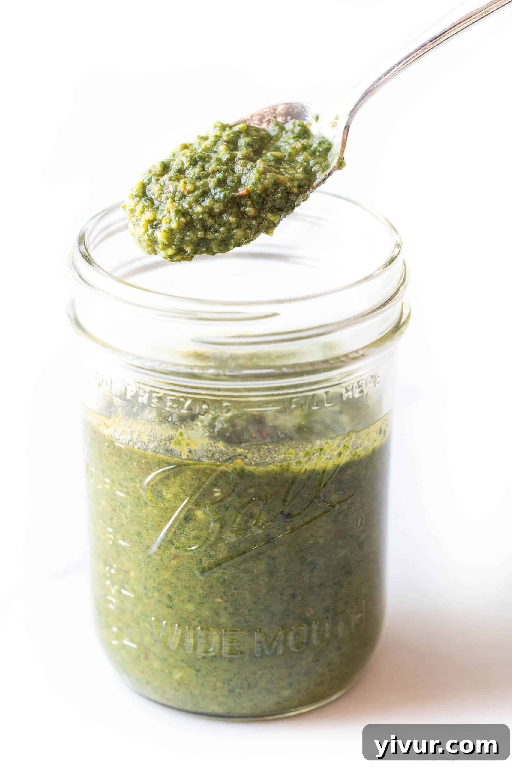 Spoon scooping vibrant green dairy-free pesto from a mason jar, showcasing its creamy texture on a pristine white surface.