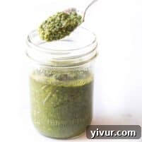 A spoon is lifting a portion of green pesto sauce from a glass jar. The jar, with Ball and Wide Mouth embossed on it, is filled with vibrant green pesto, sitting on a white surface.