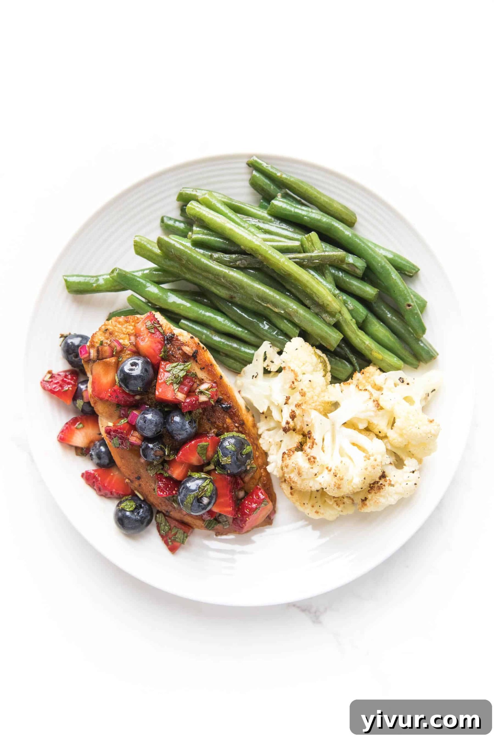 chicken topped with blueberry and strawberry salsa on a white plate and a white background with green beans and cauliflower