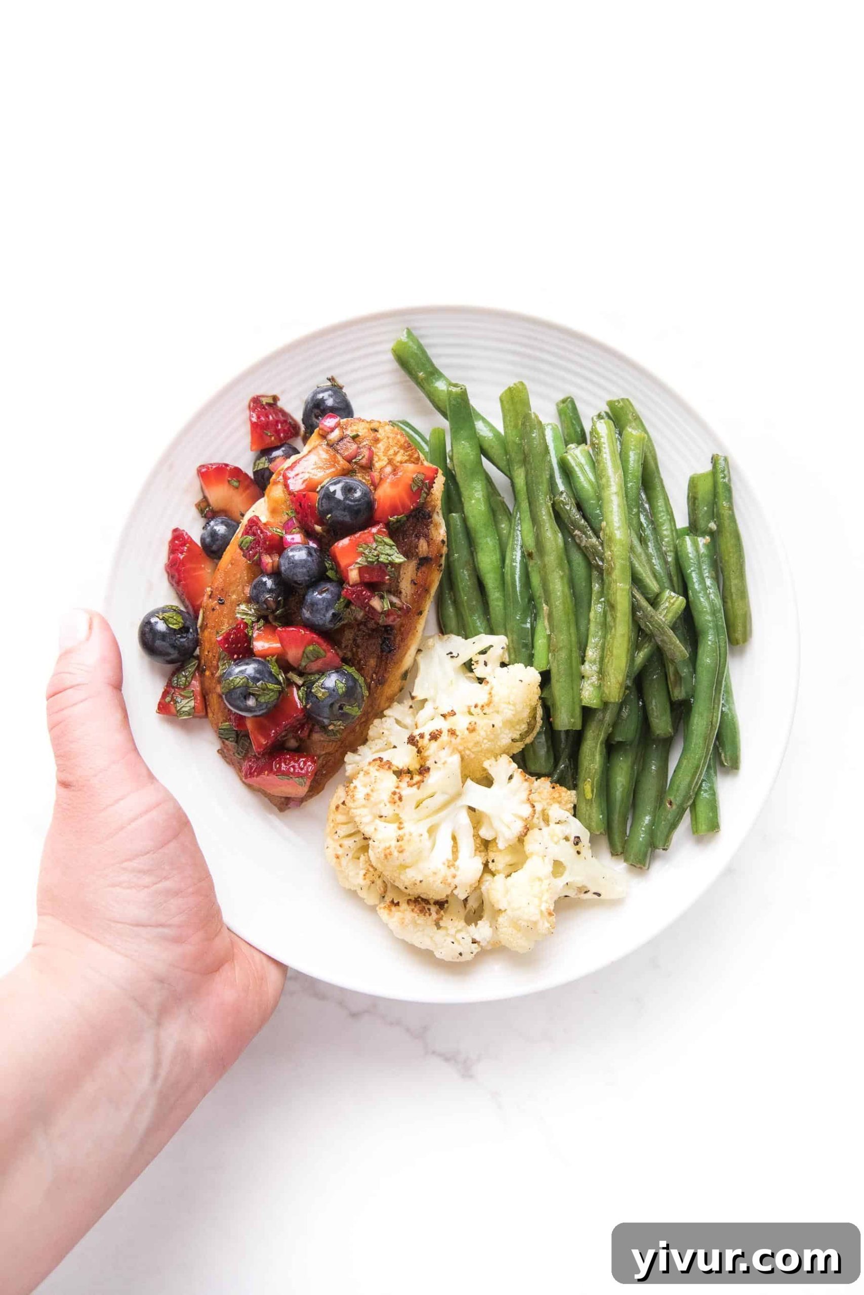 a hand holding chicken topped with blueberry and strawberry salsa on a white plate and a white background