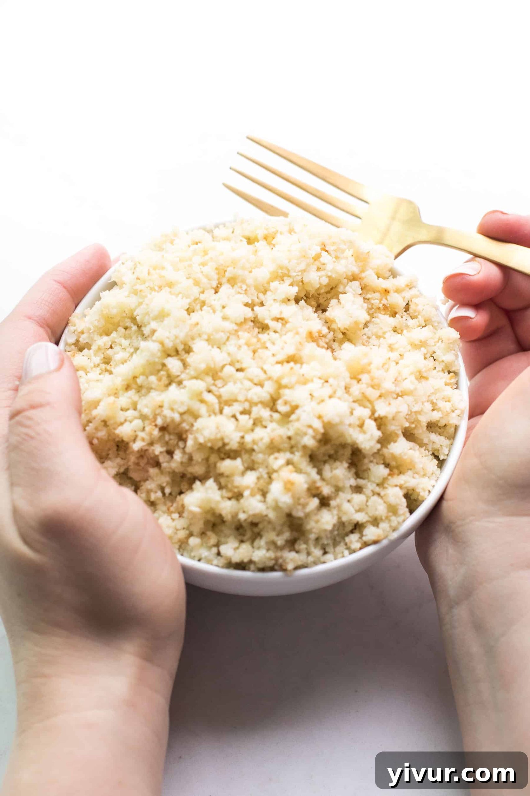 Hands gently scooping freshly cooked, fluffy homemade cauliflower rice from a white bowl with a fork, emphasizing its delightful texture.