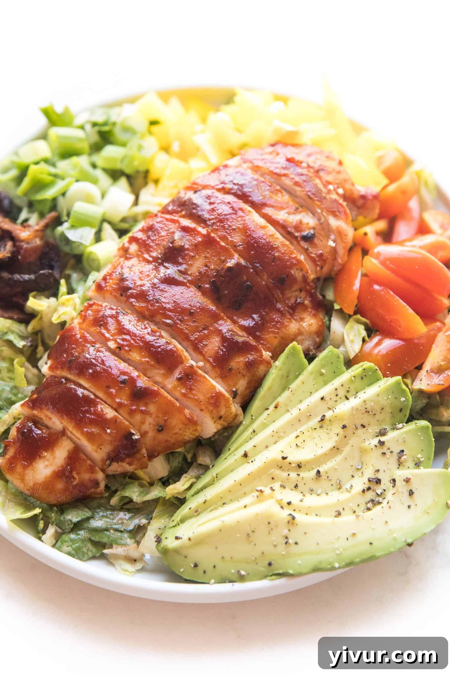 bbq chicken salad with avocado, bacon, bell pepper, tomatoes and green onions on a white plate and background
