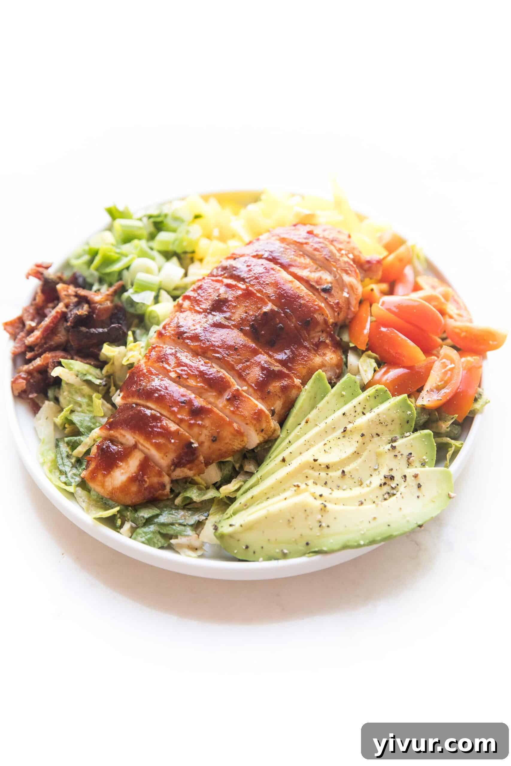 bbq chicken salad with avocado, bacon, bell pepper, tomatoes and green onions on a white plate and background