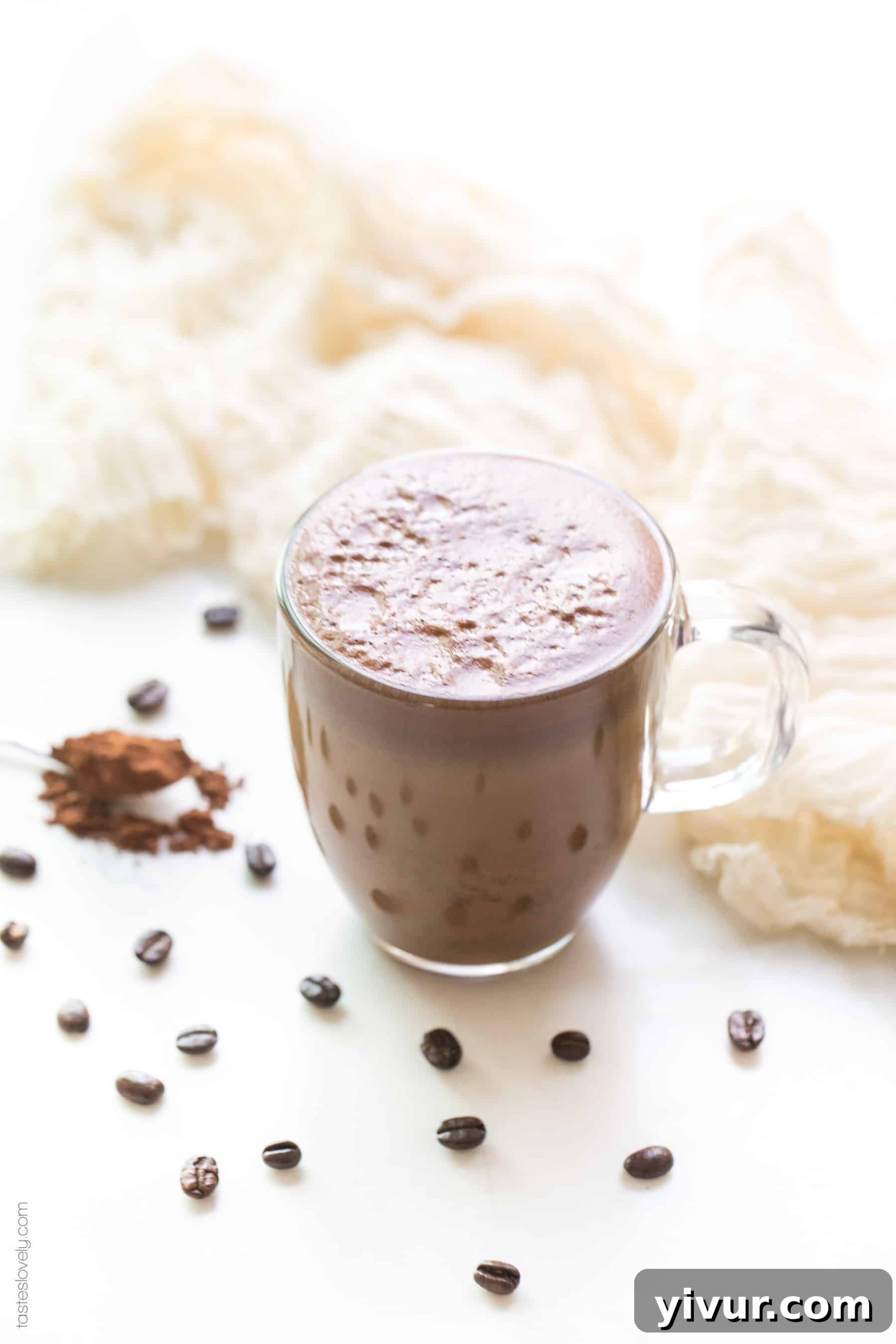 A mocha in a clear glass coffee cup on a white background with coffee beans and cocoa powder in the background, showcasing its rich texture