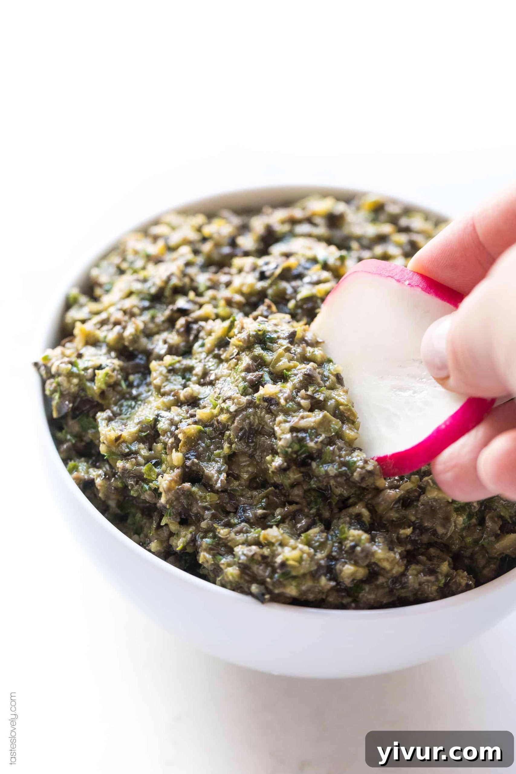 A crisp radish being dipped into a creamy white bowl of homemade olive tapenade, highlighting its fresh appeal and vibrant green garnish.
