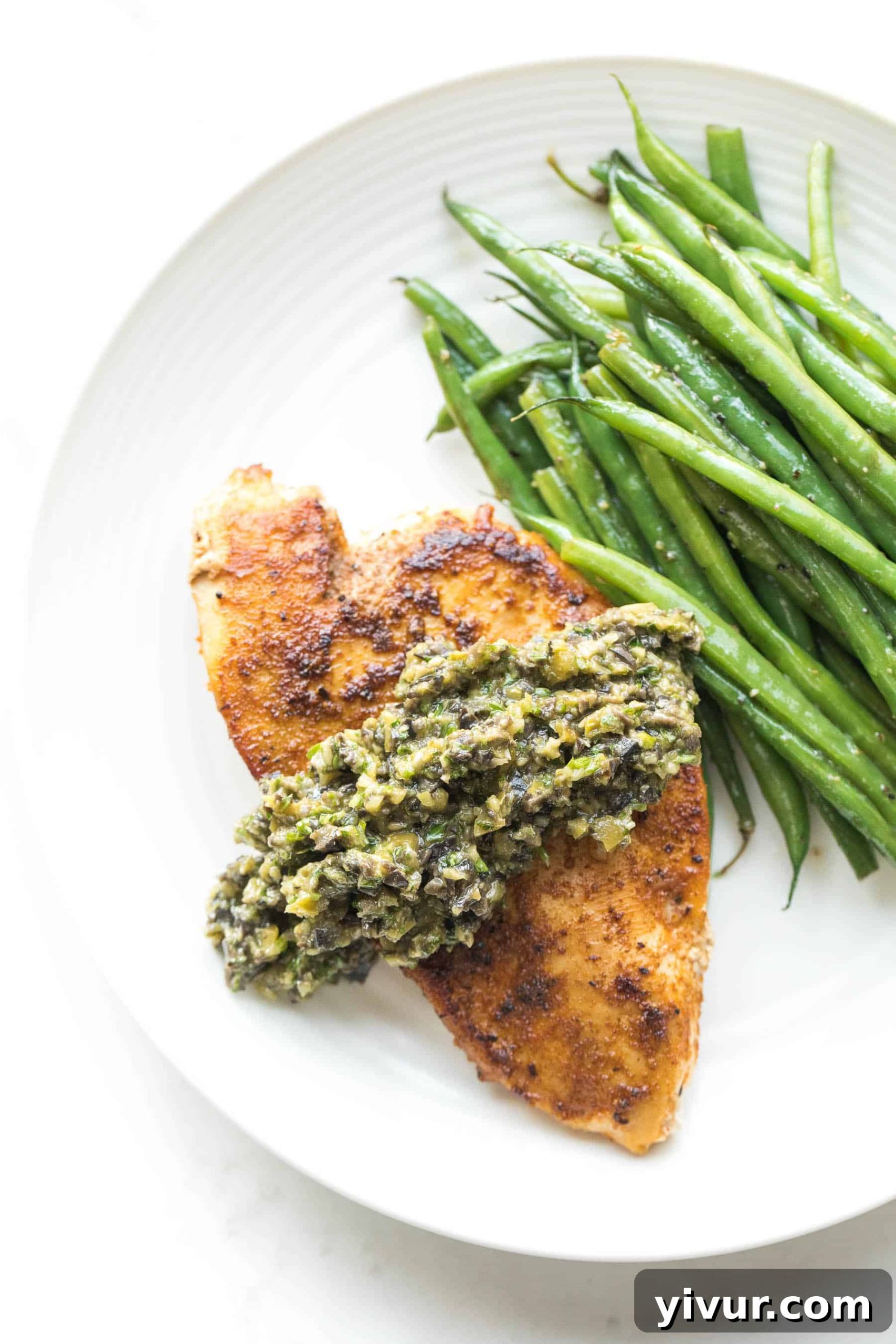 Chicken with a generous topping of olive tapenade and a side of vibrant green beans, presented elegantly on a white plate, ready to be served.