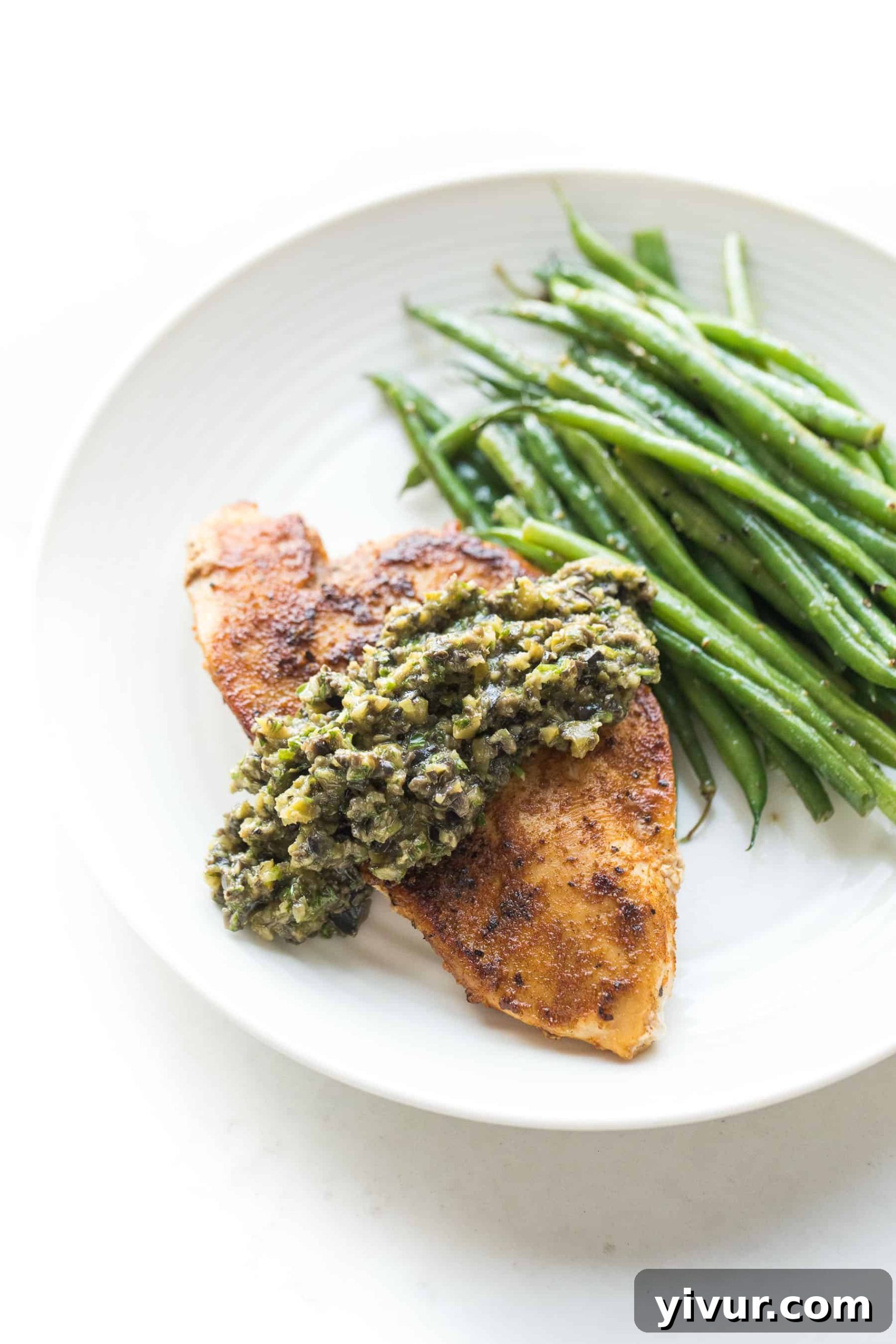 Close-up of a chicken breast topped with a generous spoonful of olive tapenade, garnished with fresh herbs and served alongside green beans on a white plate.