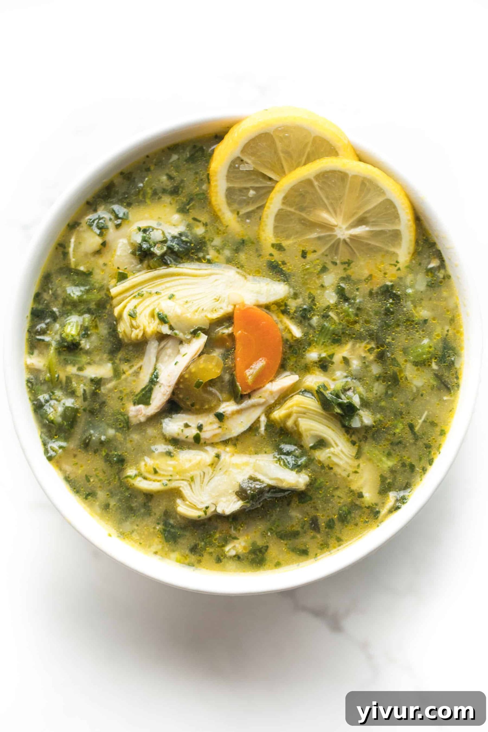 Zesty Lemon Artichoke Chicken Soup Whole30 Keto 2 A serving of Whole30 + Keto Lemon Artichoke Chicken Soup in a white bowl, garnished with two lemon slices, highlighting its fresh and inviting appearance.