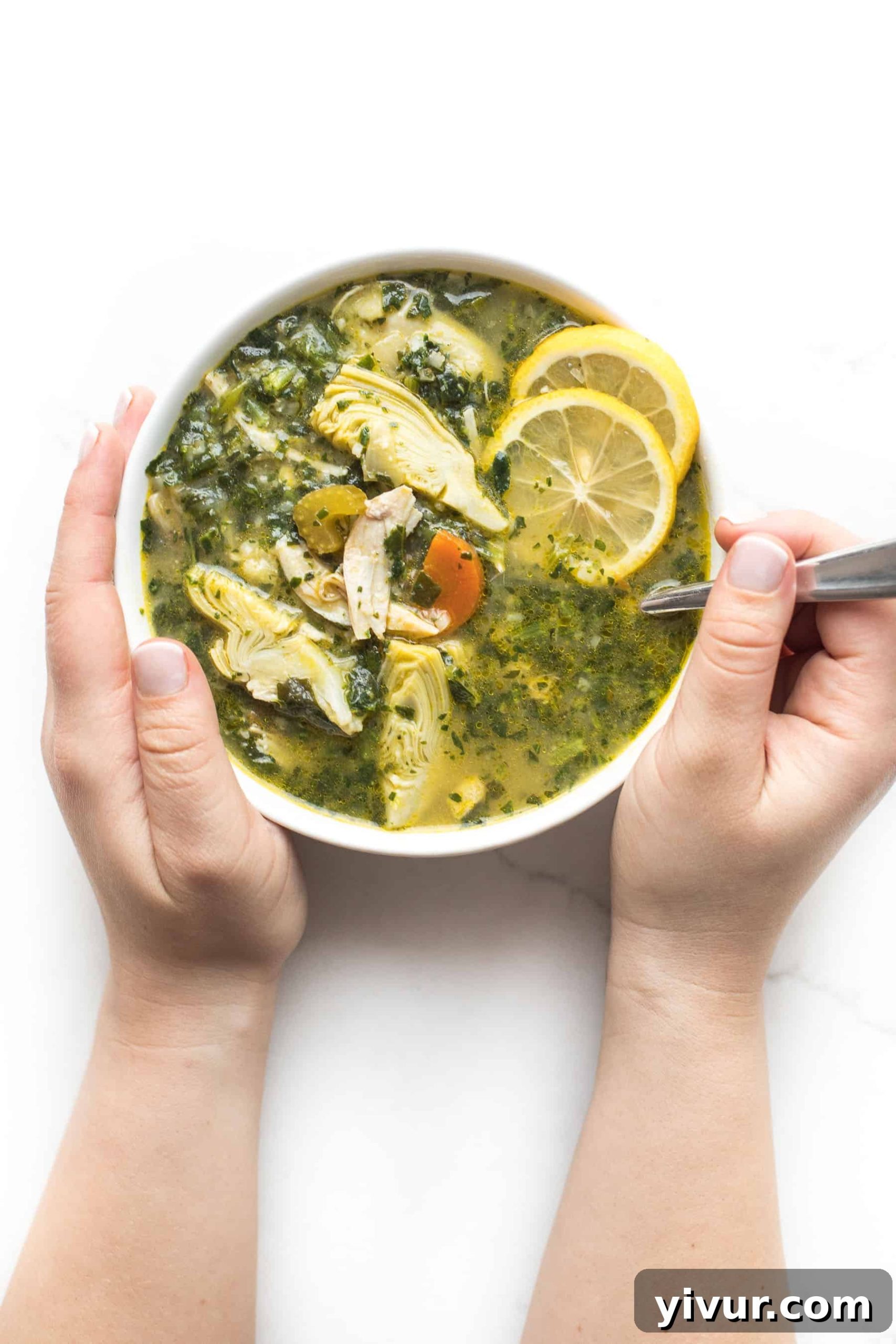 Zesty Lemon Artichoke Chicken Soup Whole30 Keto 5 Two hands gently holding a white bowl filled with the vibrant Lemon Artichoke Chicken Soup, inviting the viewer to savor its warmth and freshness.