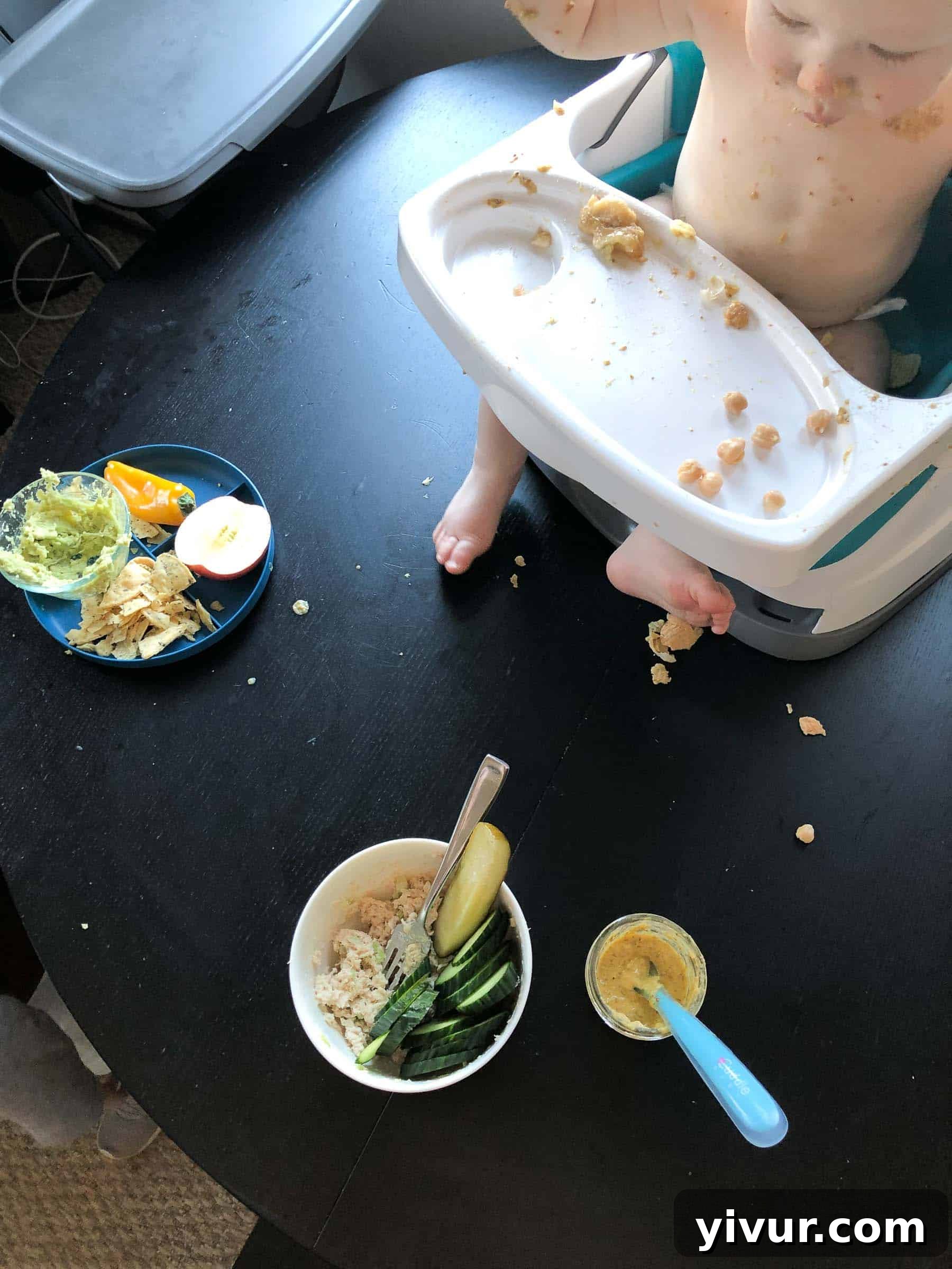 A glimpse into efficient parenting: Baby Ryan contentedly enjoying snacks in his high chair, featuring chickpeas and banana, while his mom adeptly prepares lunch, demonstrating skillful multitasking in a busy household.