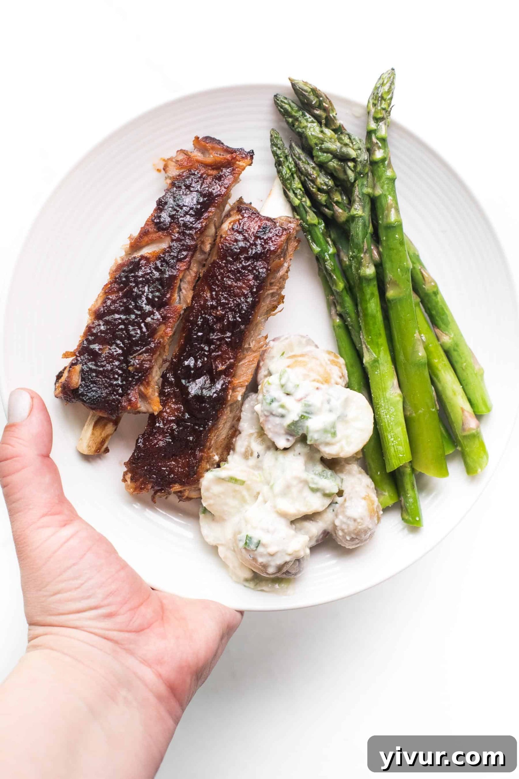 A hand holding a white plate with BBQ ribs, asparagus, and a scoop of potato salad