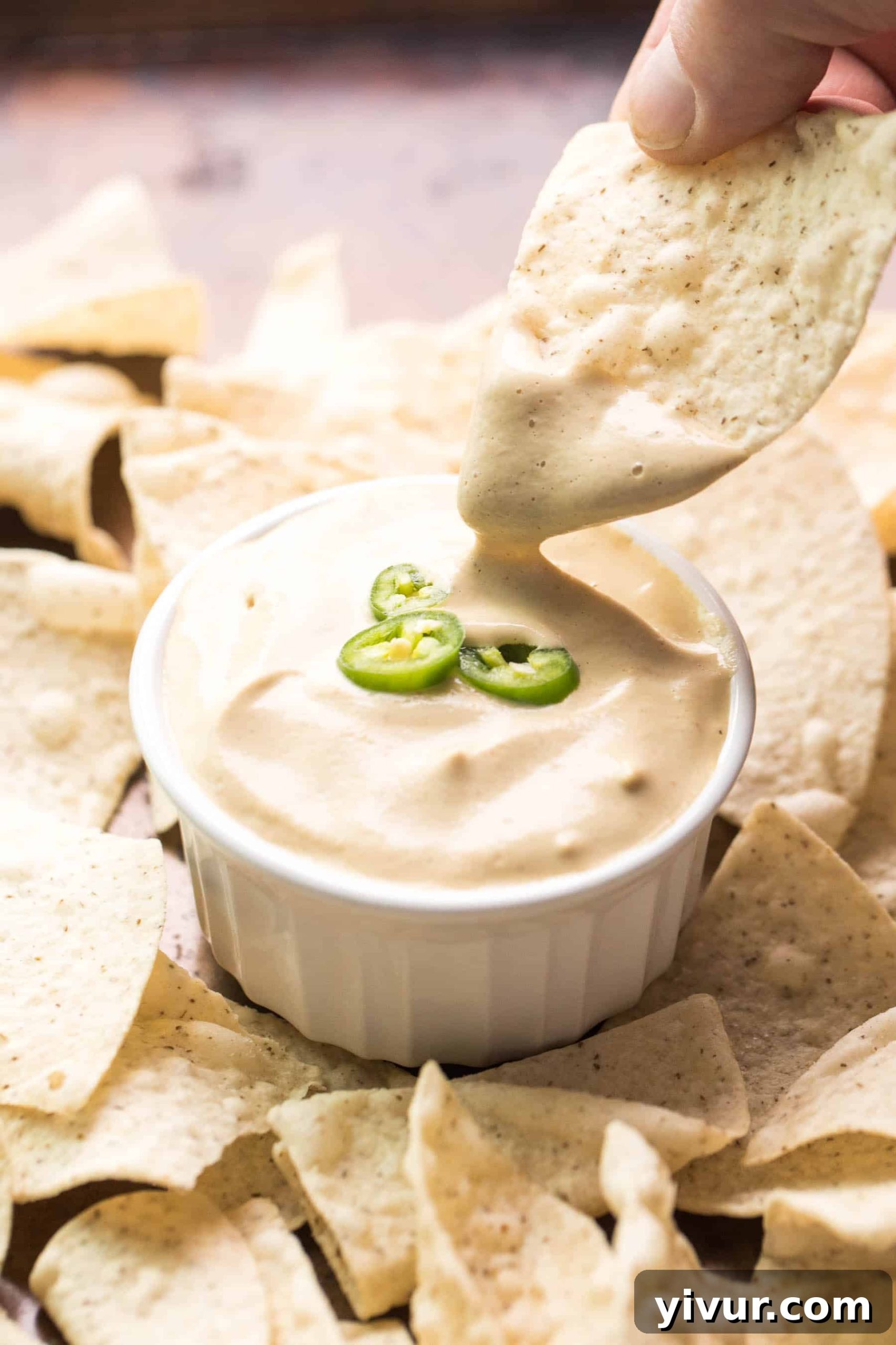 A tortilla chip being dipped into a pristine white bowl of creamy Whole30 Dairy-Free Queso Nacho Cheese, highlighting its smooth texture and appetizing color.