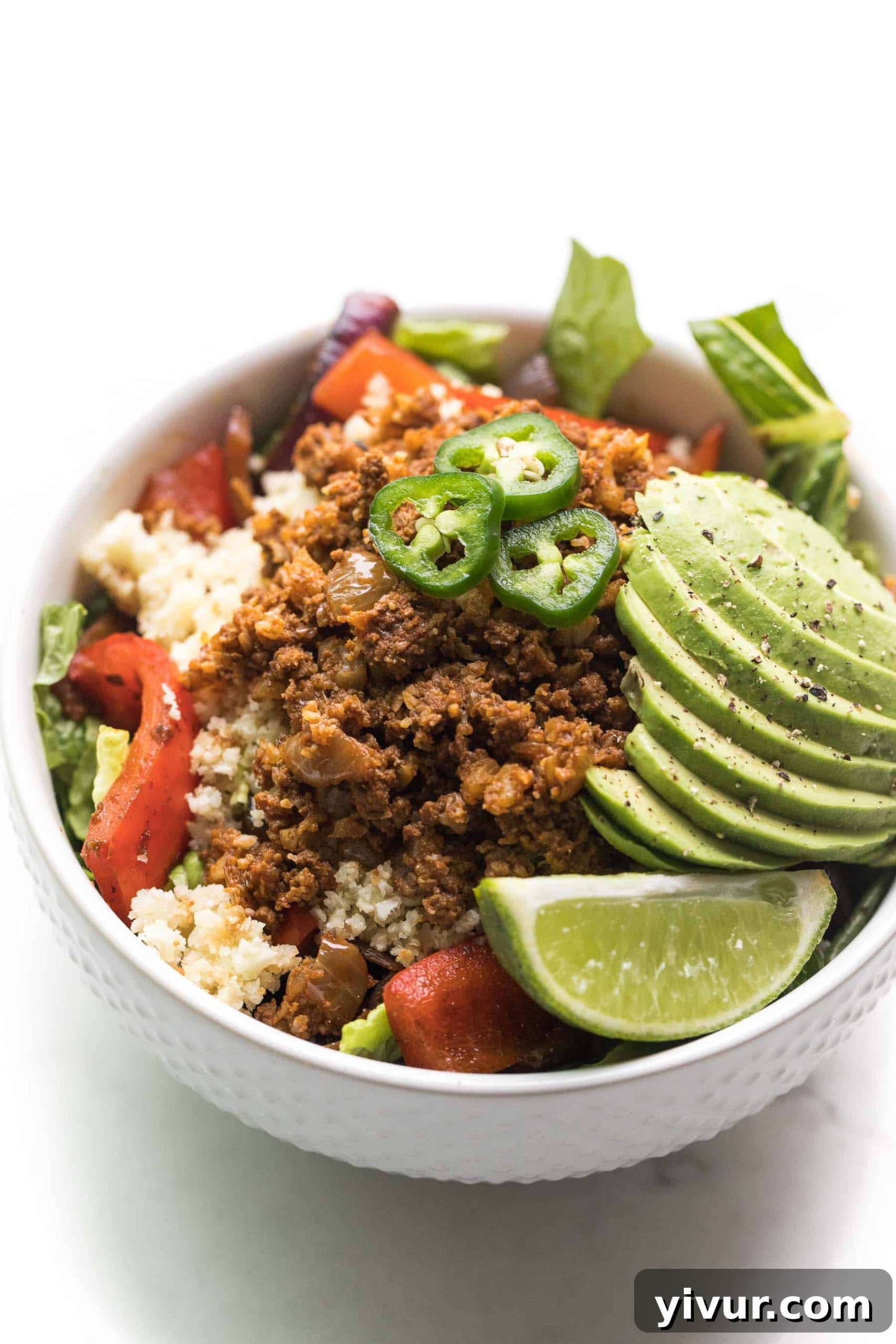 white bowl filled with ground beef, cauliflower rice, veggies, and lettuce, and topped with jalapeno, avocado, and lime