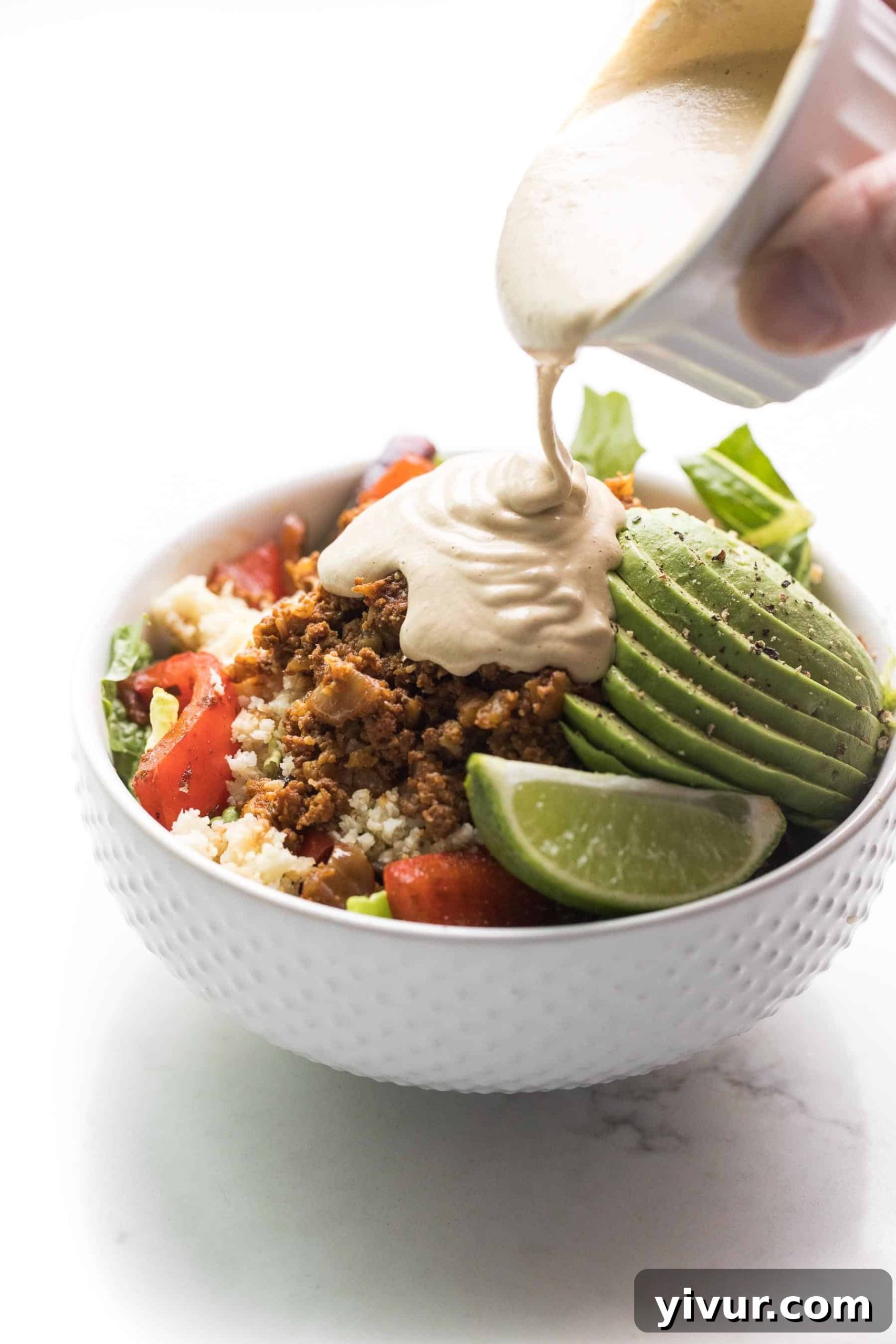 a hand pouring queso over a beef burrito bowl in a white bowl