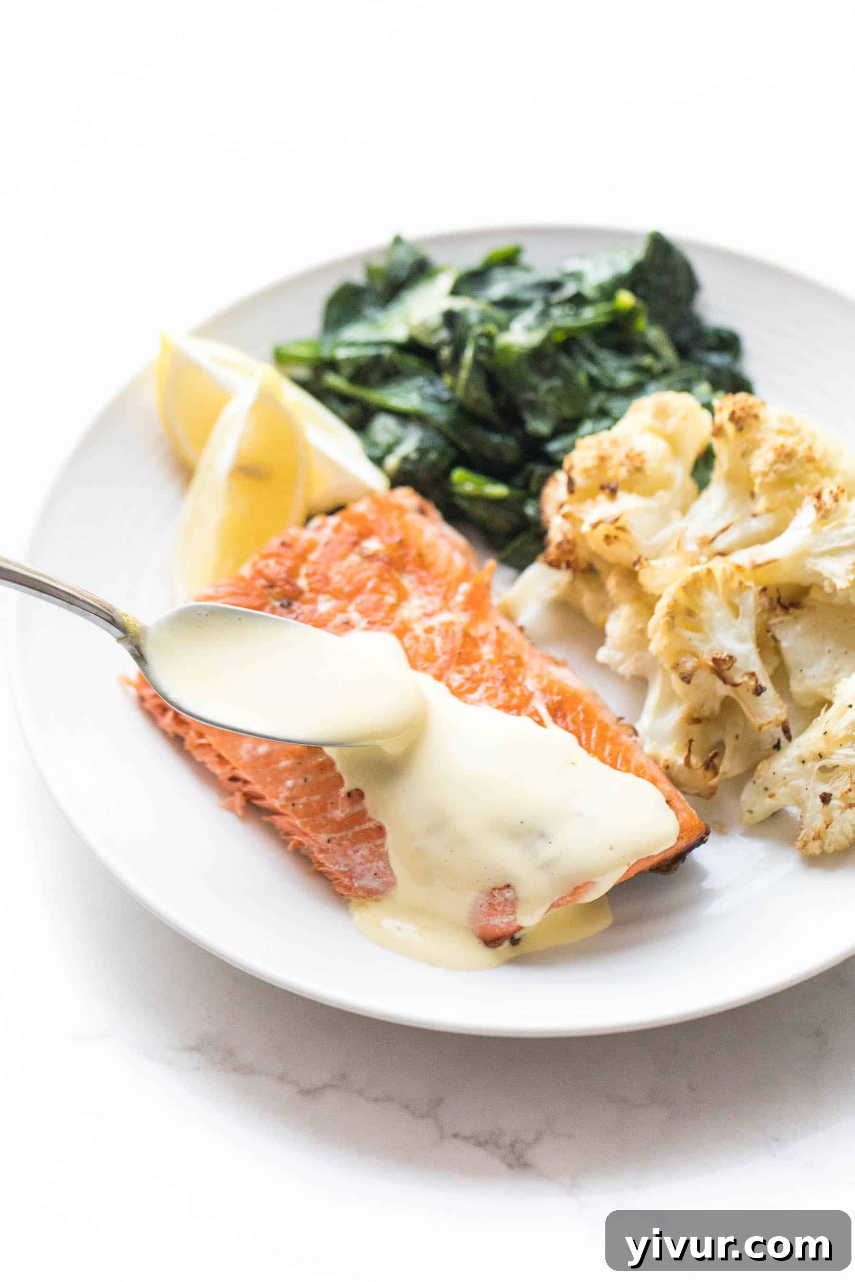 White plate with Whole30 salmon and a spoon pouring hollandaise sauce, roasted cauliflower, and sauteed spinach on a white background.