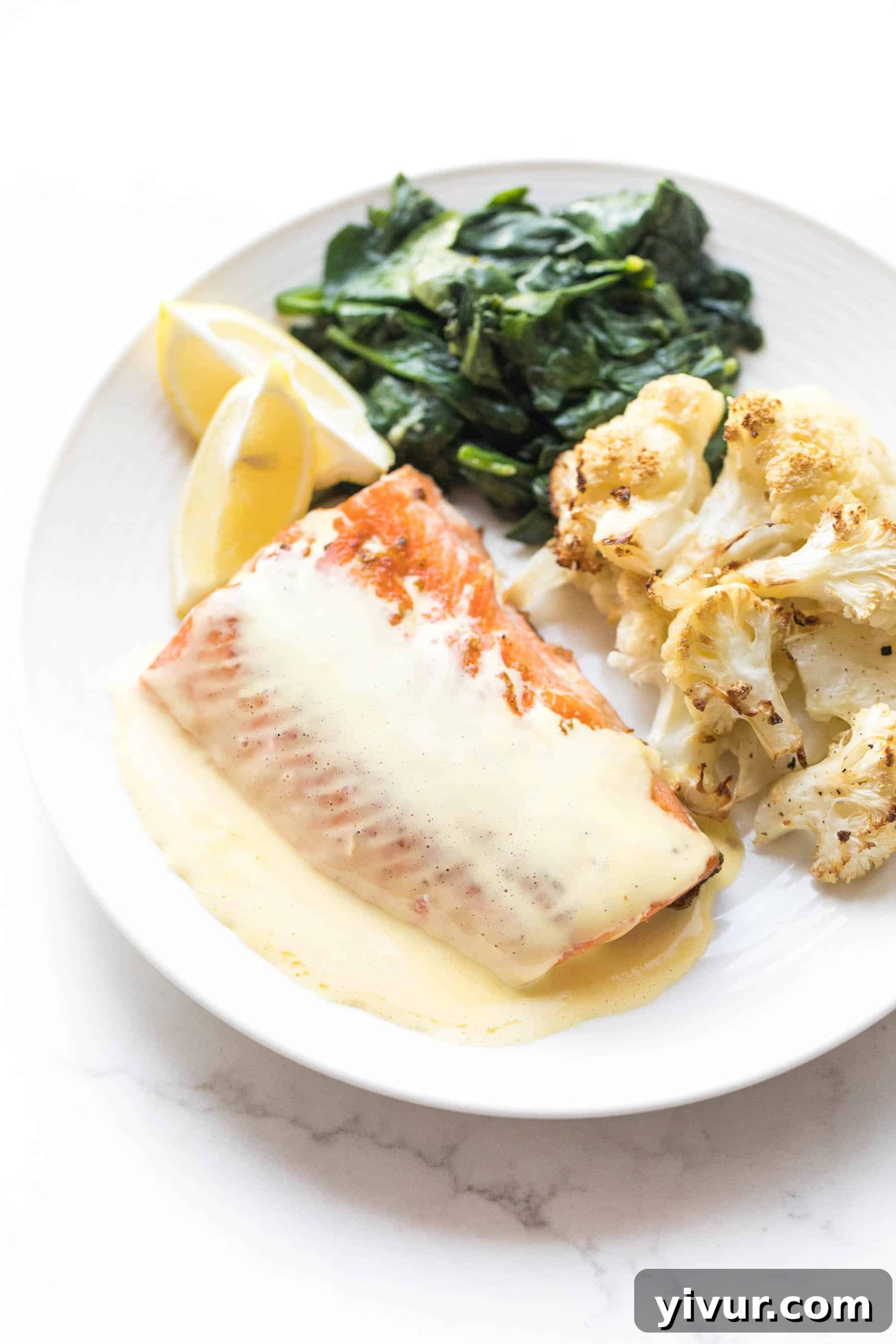 Whole30 salmon recipe on a white plate and hollandaise sauce, roasted cauliflower, lemon wedges, and sauteed spinach on a white background.