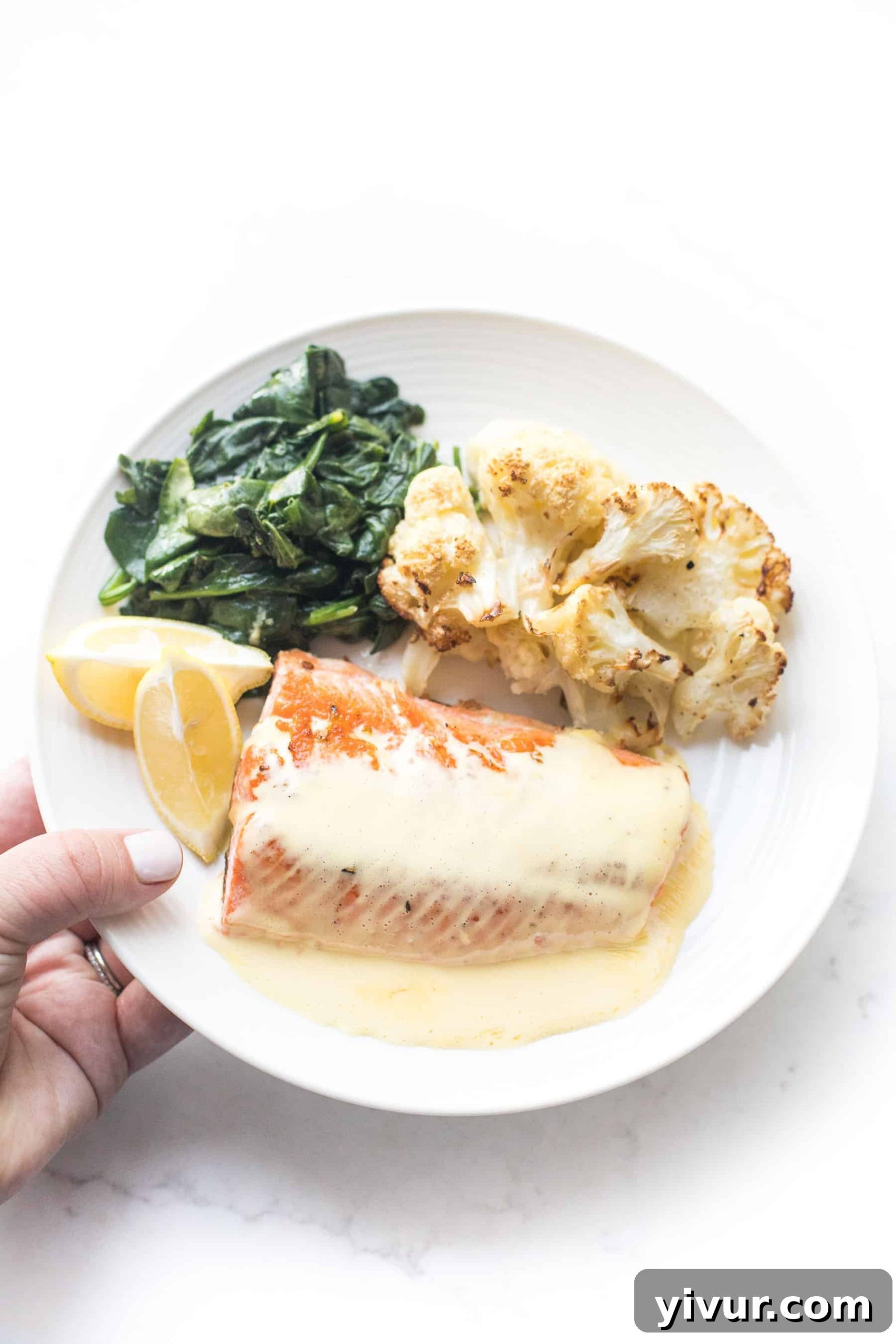 A hand holding a white plate with salmon and hollandaise sauce, roasted cauliflower, sauteed spinach, and lemon wedges on a white background.