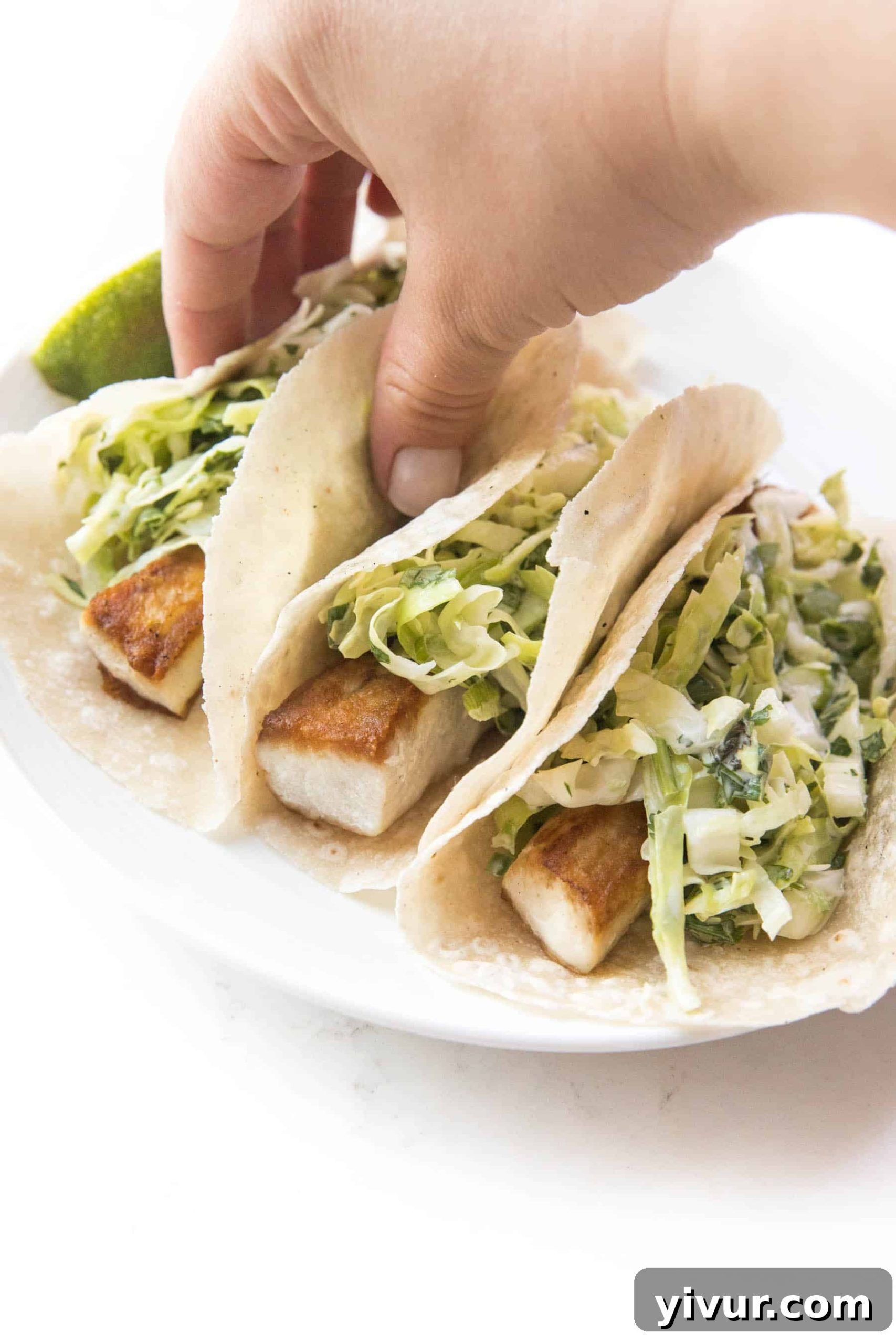 Whole30 Keto Fish Tacos with Fiery Slaw 5 A hand reaches for a Whole30 Keto Fish Taco, highlighting the generous topping of spicy coleslaw and fresh avocado slices.
