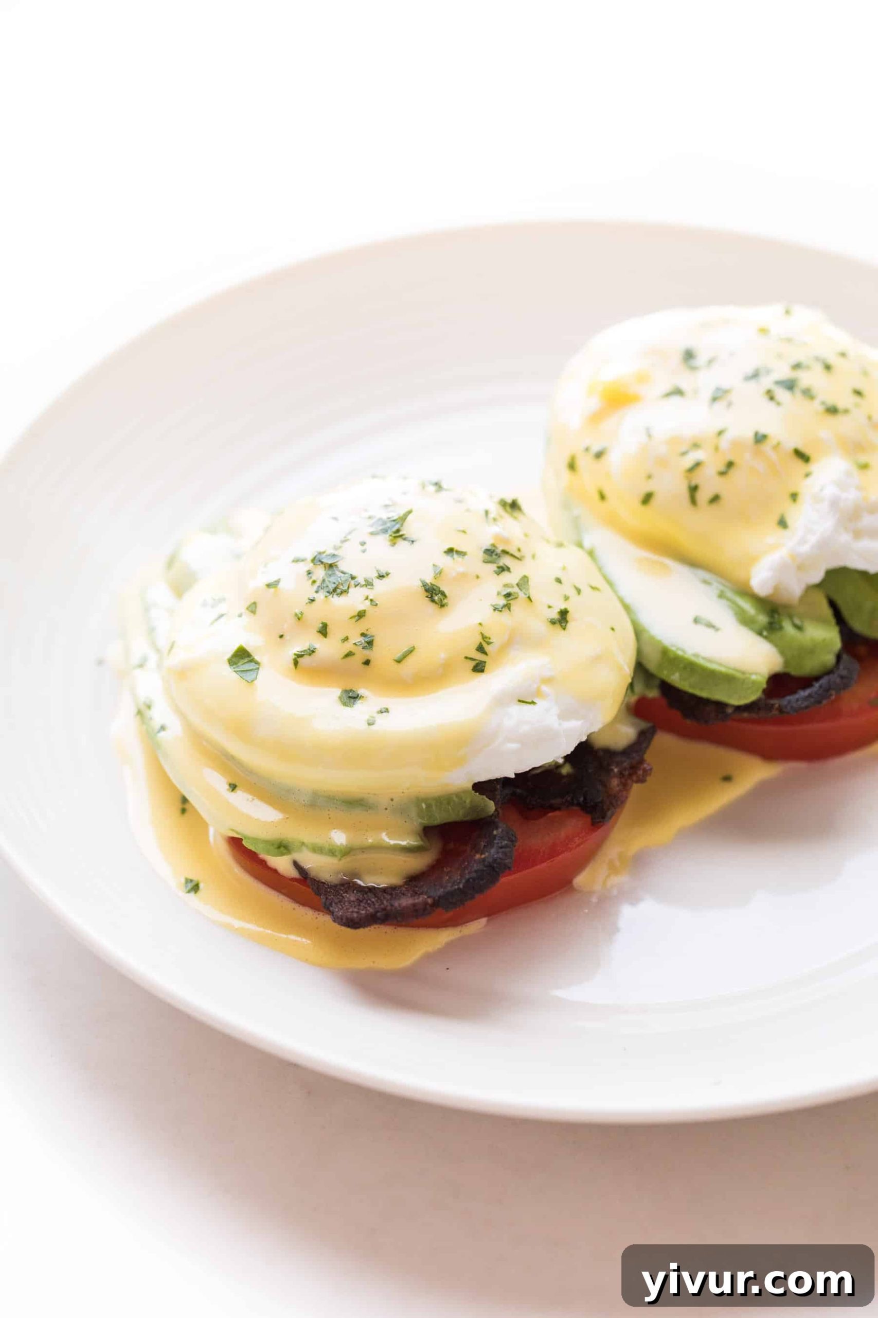 Whole30 and keto-friendly eggs benedict stacked with avocado, bacon, poached eggs, and creamy hollandaise on a white plate.