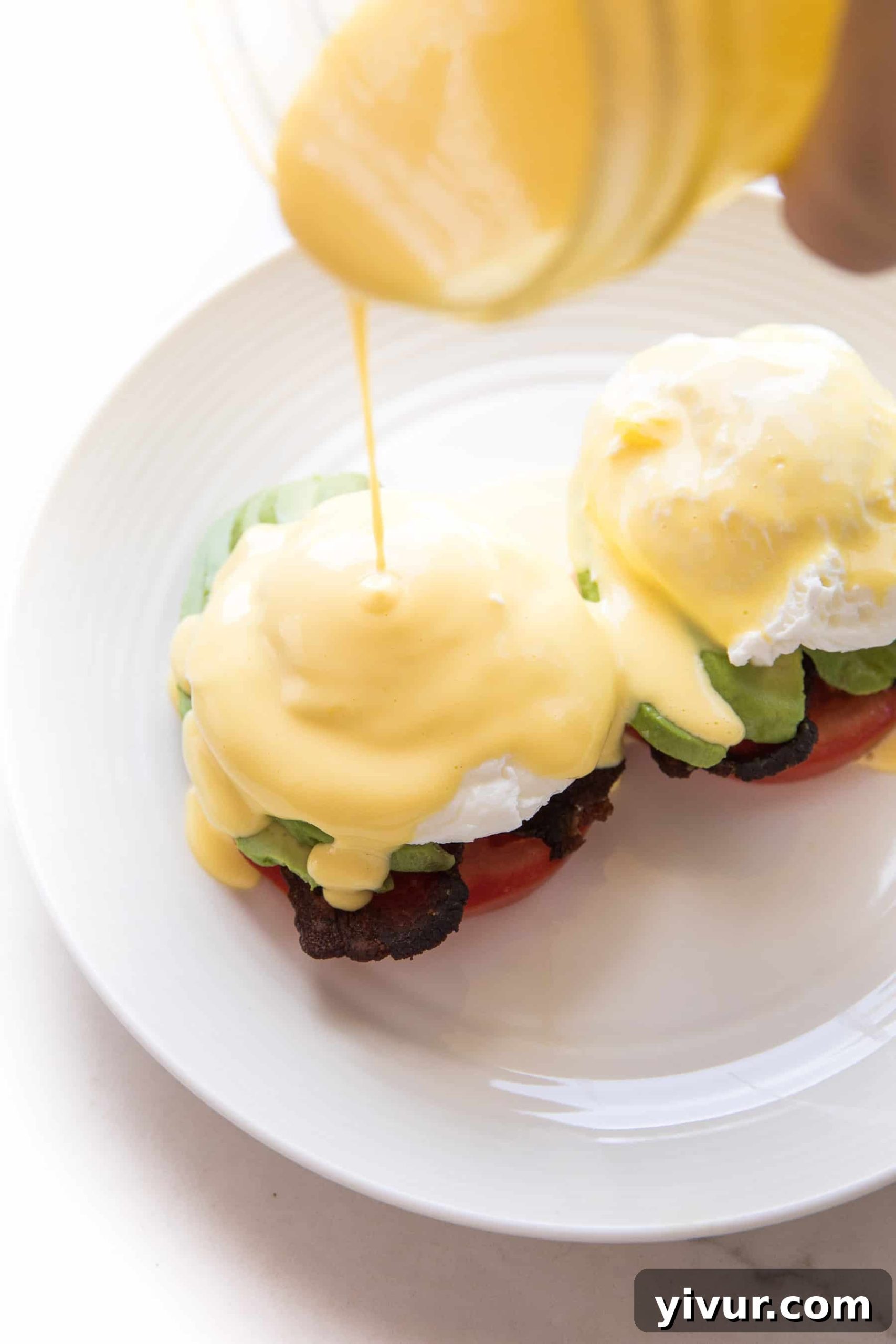 Hollandaise sauce being poured over a stack of keto eggs benedict with tomatoes and poached eggs on a white plate.