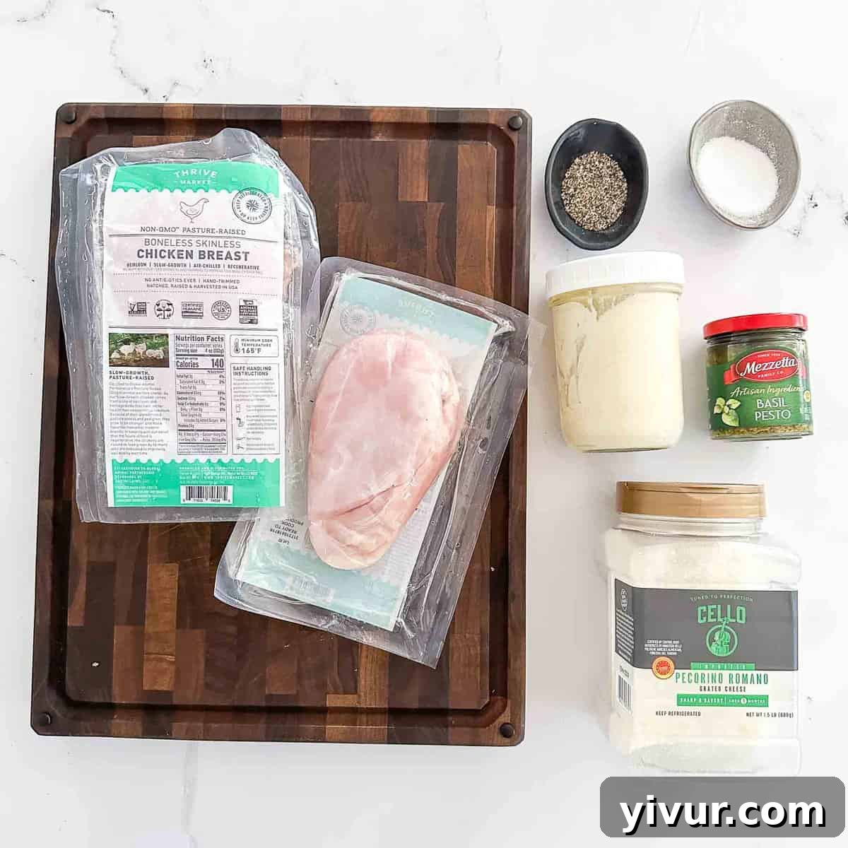 A beautiful display of fresh recipe ingredients on a rustic wooden cutting board, featuring chicken breasts, a bowl of vibrant pesto, creamy mayonnaise, and a mix of herbs and spices.