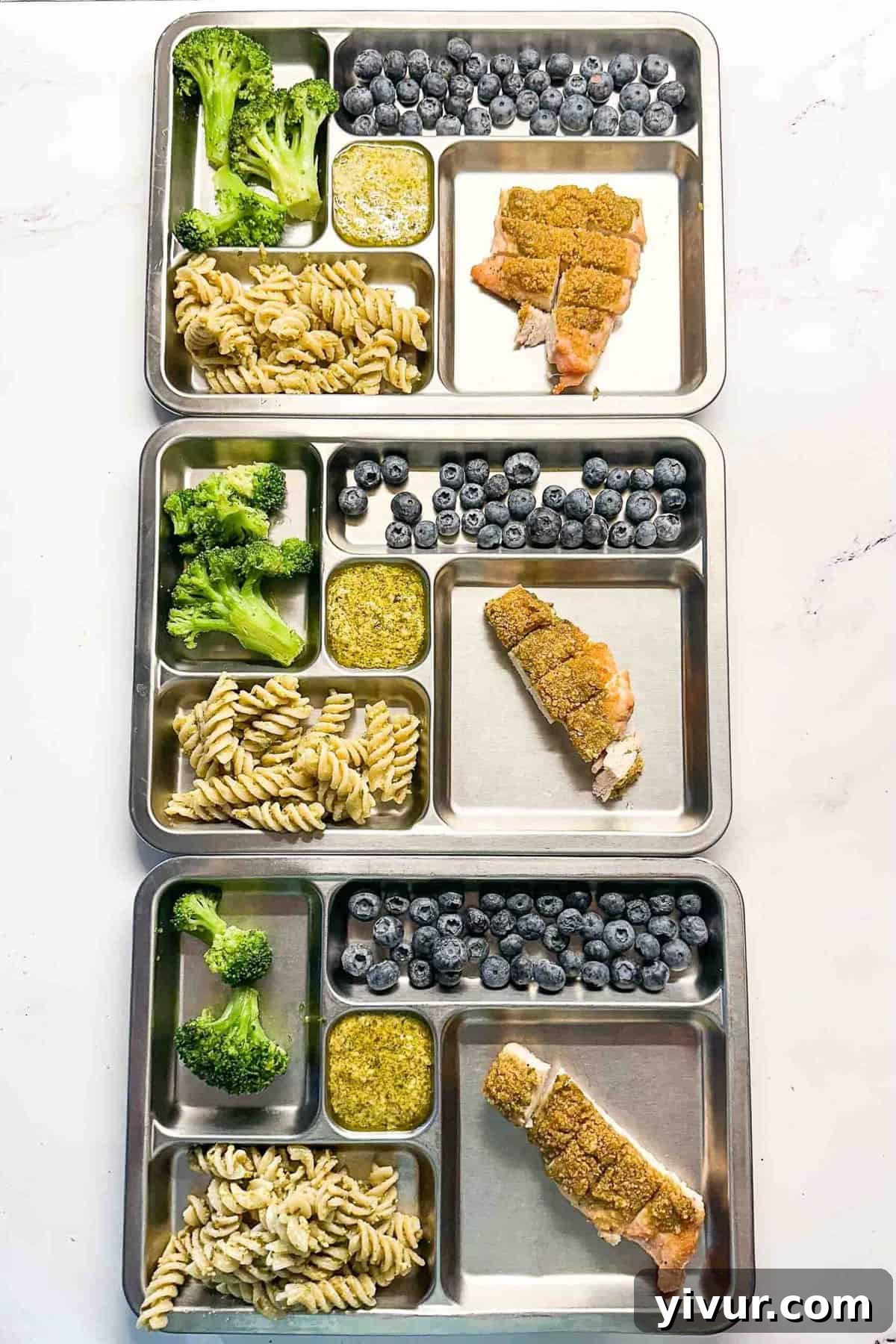 Three metal kids' plates, each featuring a portion of crispy baked pesto chicken alongside healthy sides like roasted broccoli and fruit, demonstrating kid-friendly meal presentation.