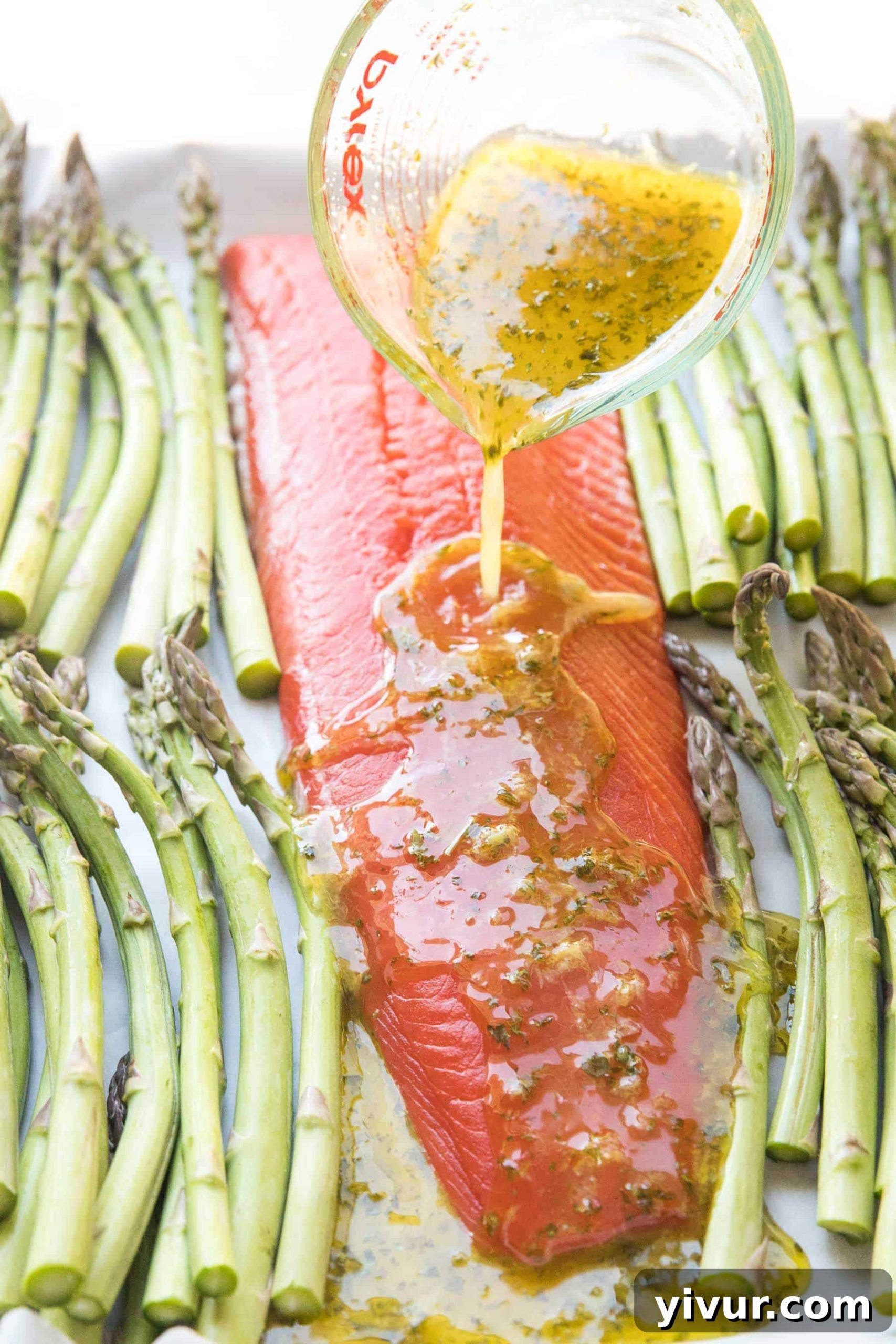 Pouring a fresh lemon-herb marinade over raw salmon and asparagus on a baking sheet