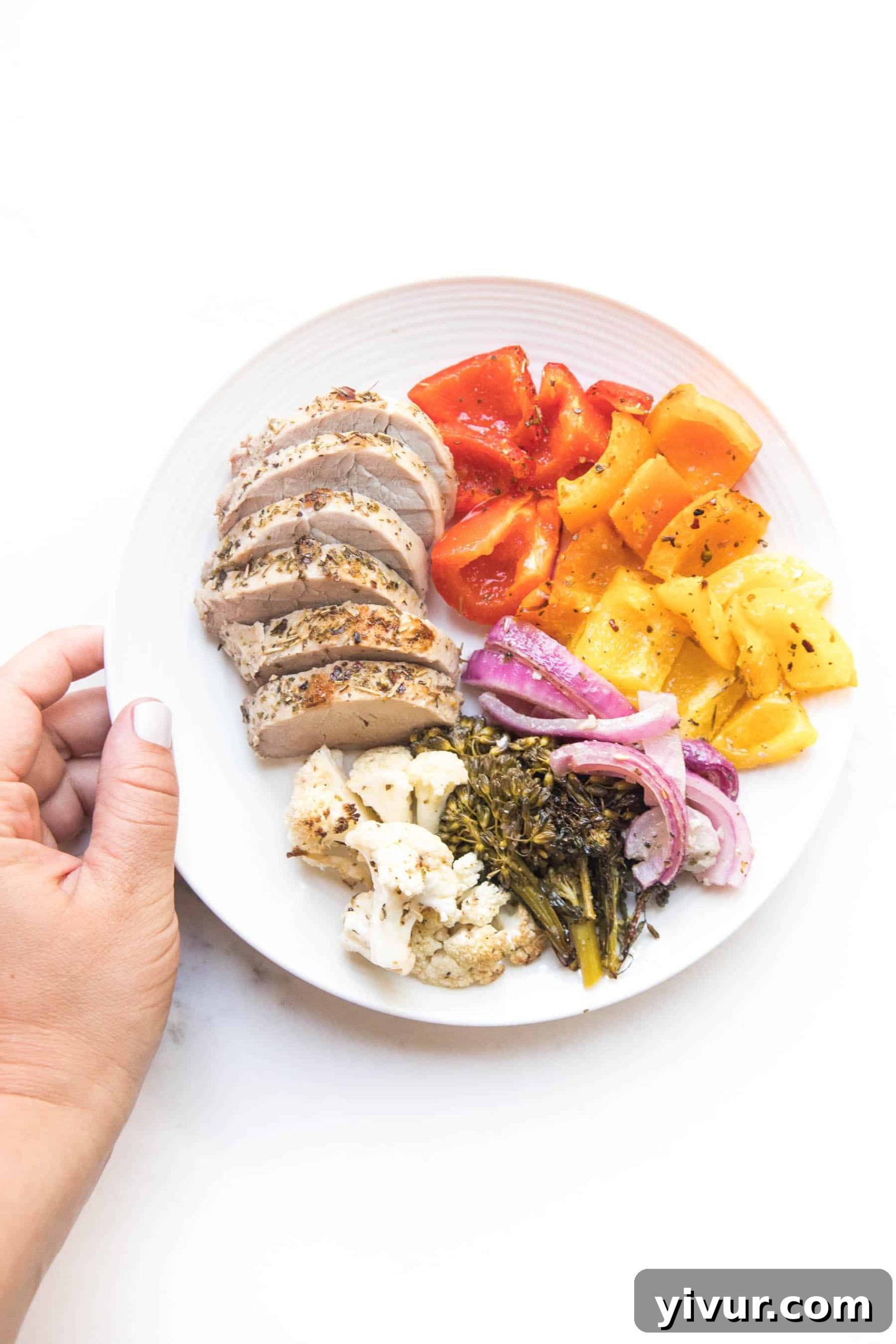 A hand holding a white plate showcasing perfectly sliced, succulent Italian pork tenderloin alongside a colorful medley of roasted vegetables
