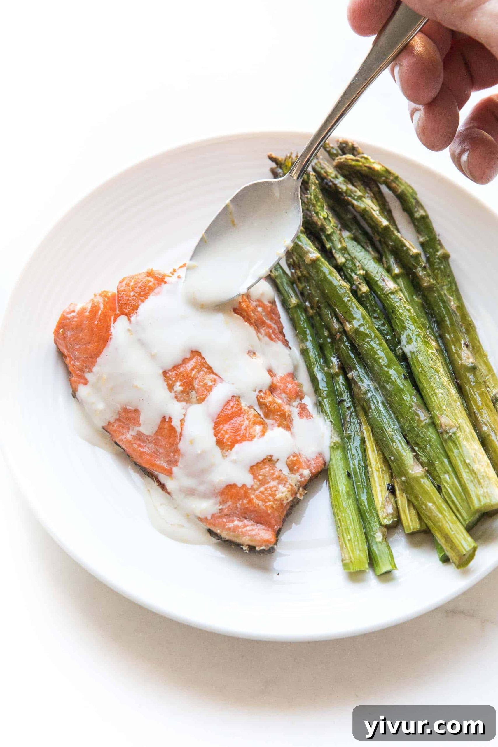 spoon drizzling Salmon + asparagus with a white creamy coconut tahini sauce on a white plate and background