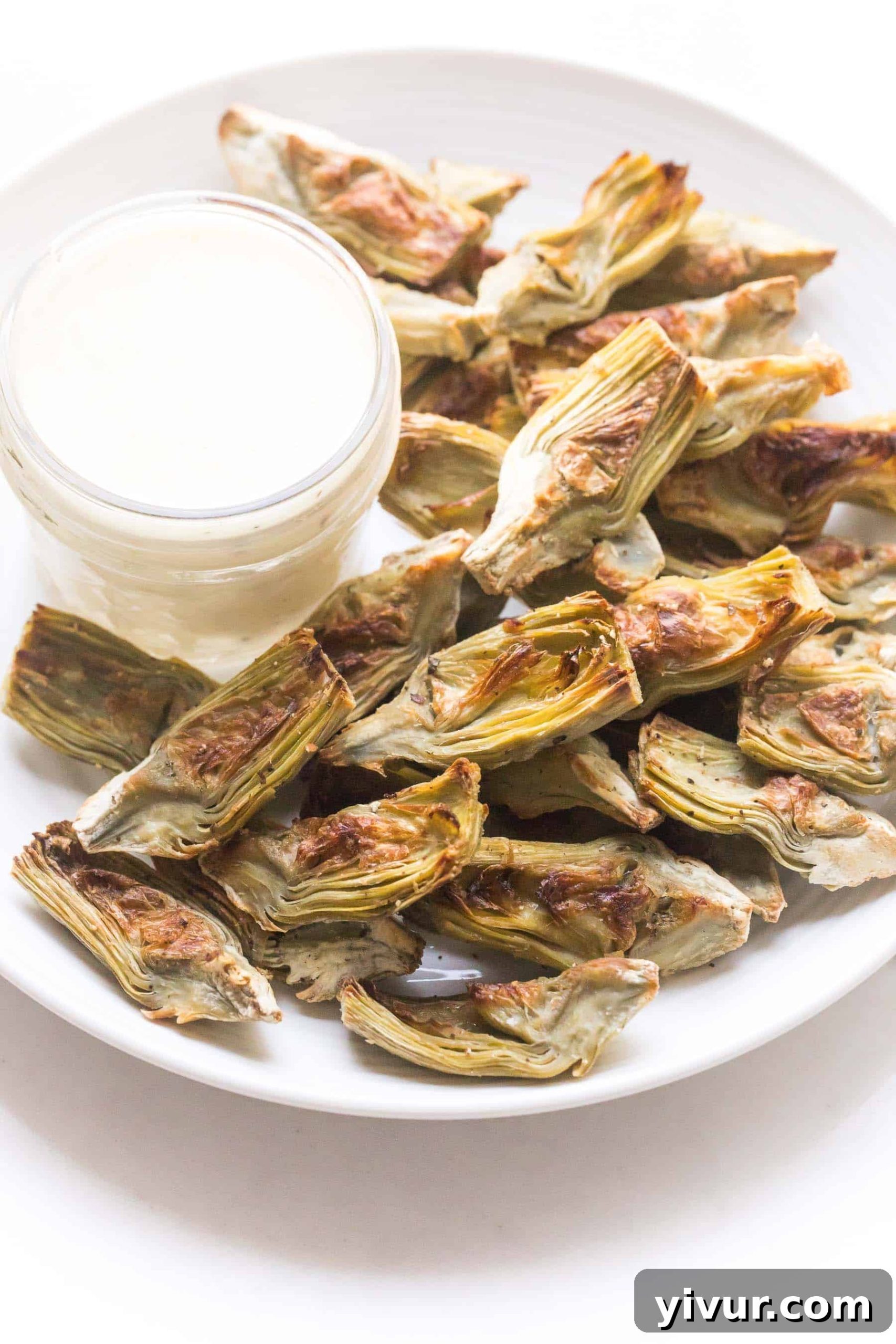Roasted artichoke hearts on a white plate with lemon garlic aioli dipping sauce on a white background