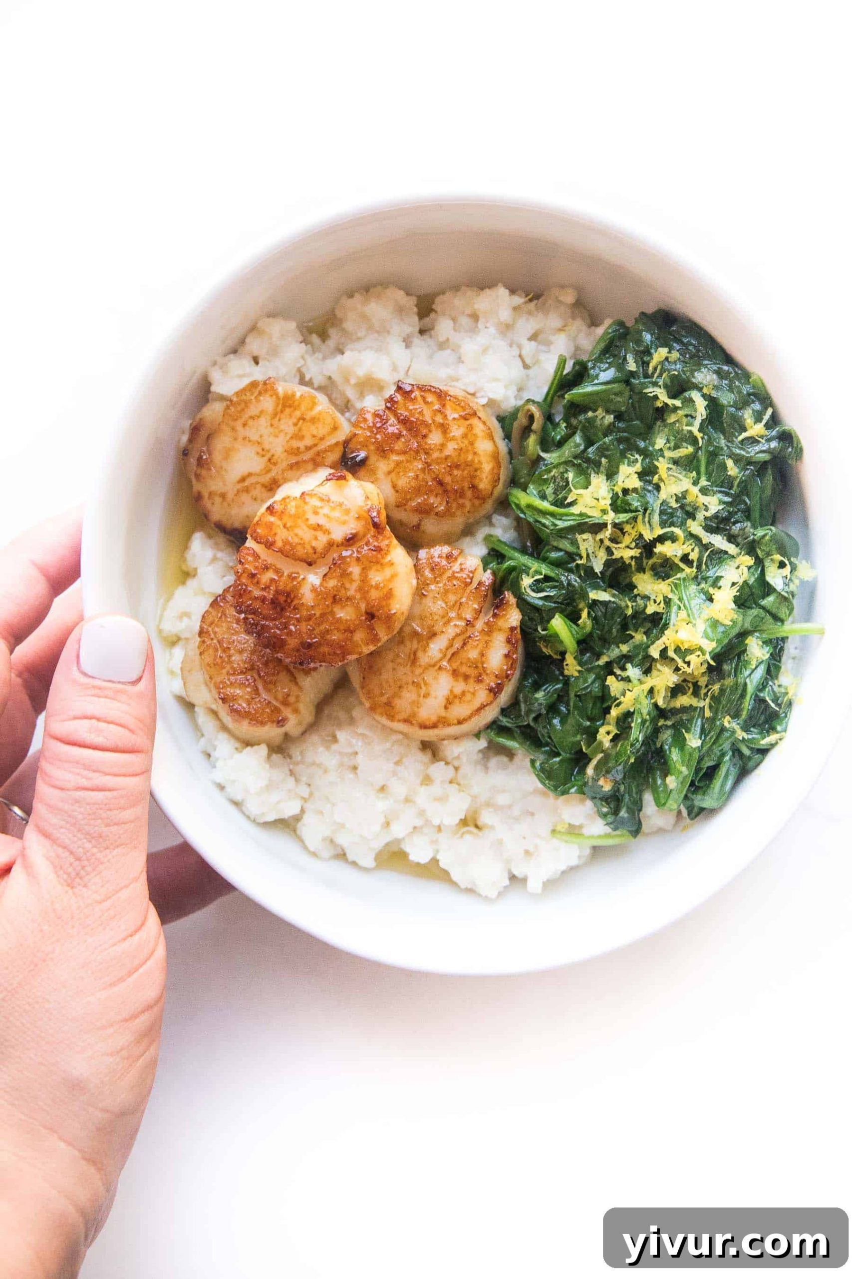 Crispy Golden Whole30 Keto Scallops 5 A hand holding a white bowl filled with perfectly seared scallops, accompanied by cauliflower rice risotto and sautéed spinach, ready to be enjoyed.