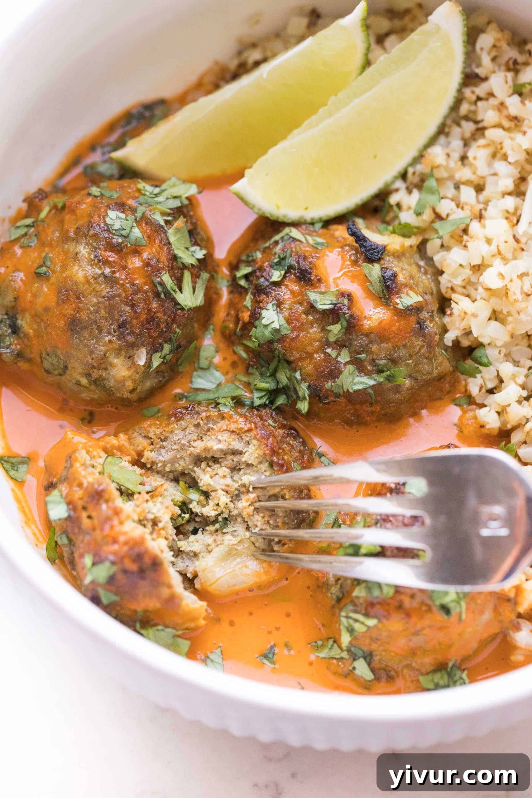 A fork cutting into a Thai curry meatball in a white bowl, showing the juicy interior, surrounded by creamy sauce and fresh herbs.