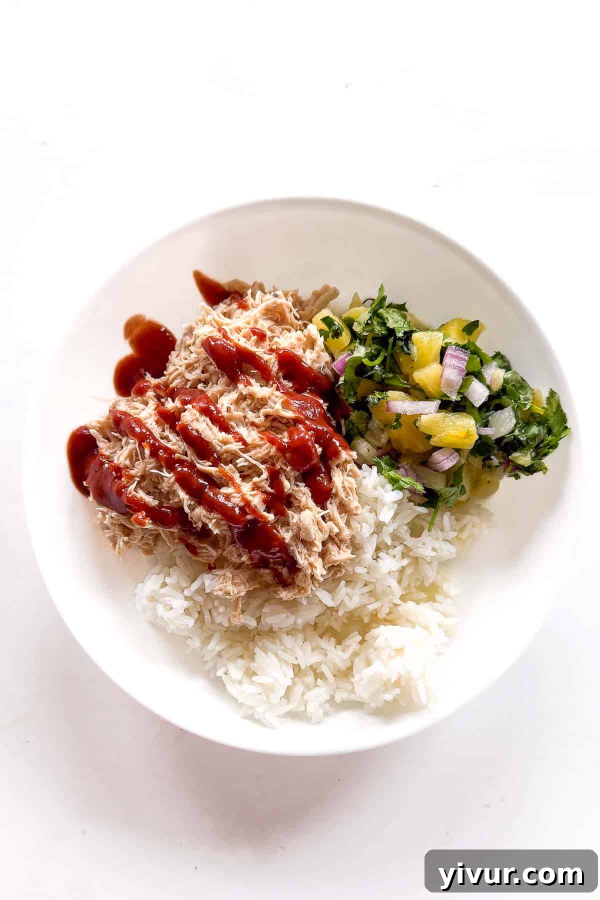 A vibrant white bowl showcases tender, high-protein shredded Hawaiian Chicken nestled atop a bed of fluffy white rice. The chicken is generously drizzled with a rich red BBQ sauce and garnished with fresh green herbs. A side of bright pineapple salsa, featuring finely chopped red onion and cilantro, adds a refreshing contrast, making for a perfectly balanced and appealing meal.
